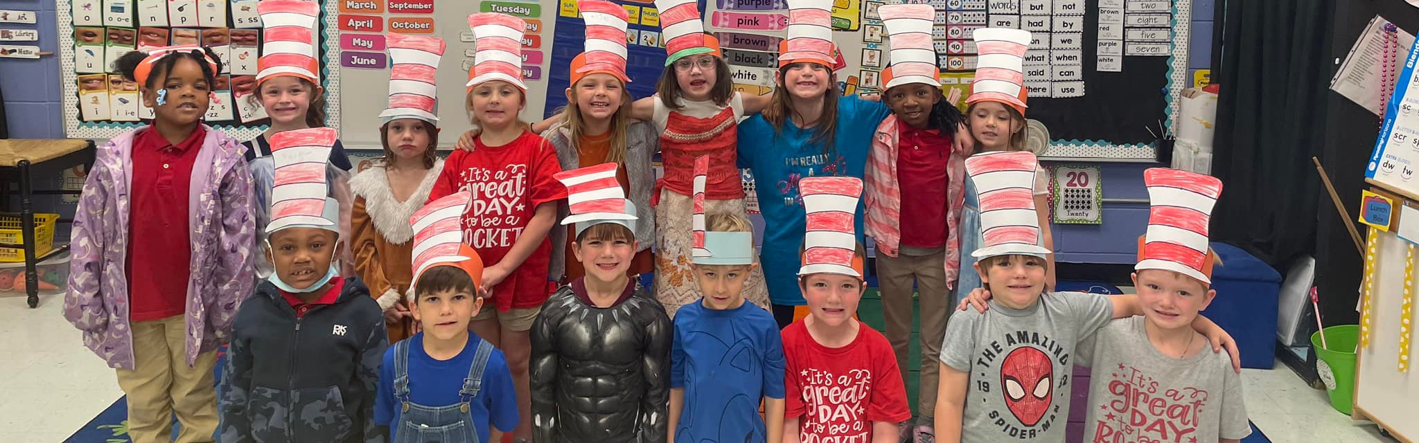 A group of about 20 elementary students wear red and white cardboard Dr. Seuss hats. They are posing in their colorful classroom