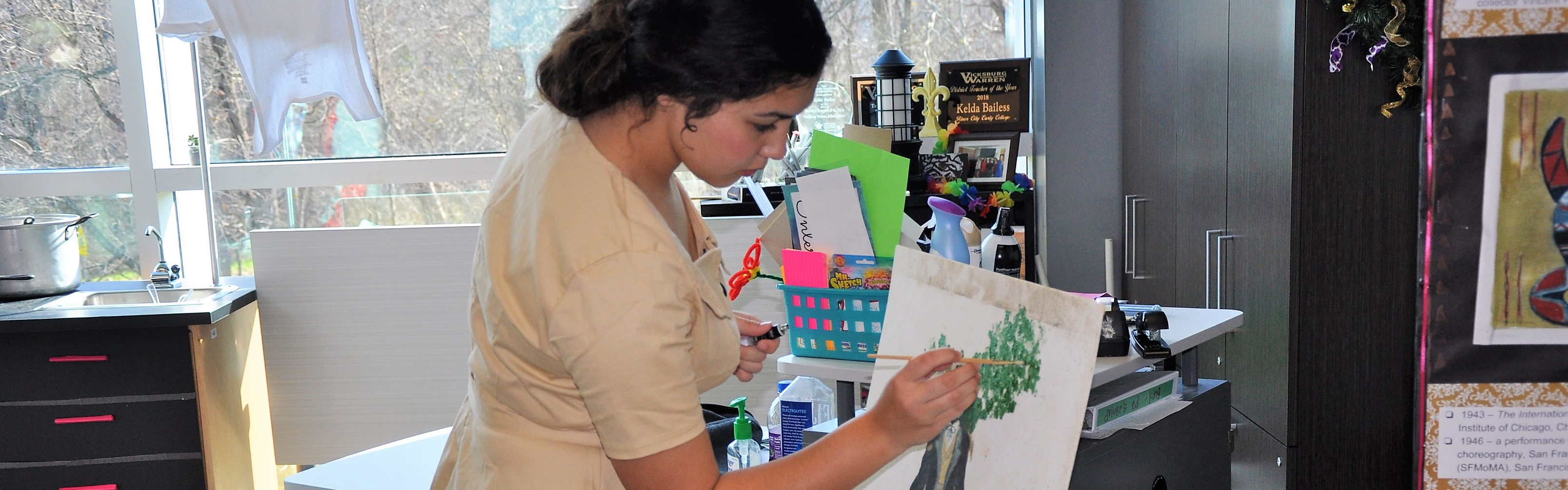 Young white female student paints at an easel. She is painting an image of a gentleman wearing a suit with a green tree in the background. The student is wearing a beige dress. 