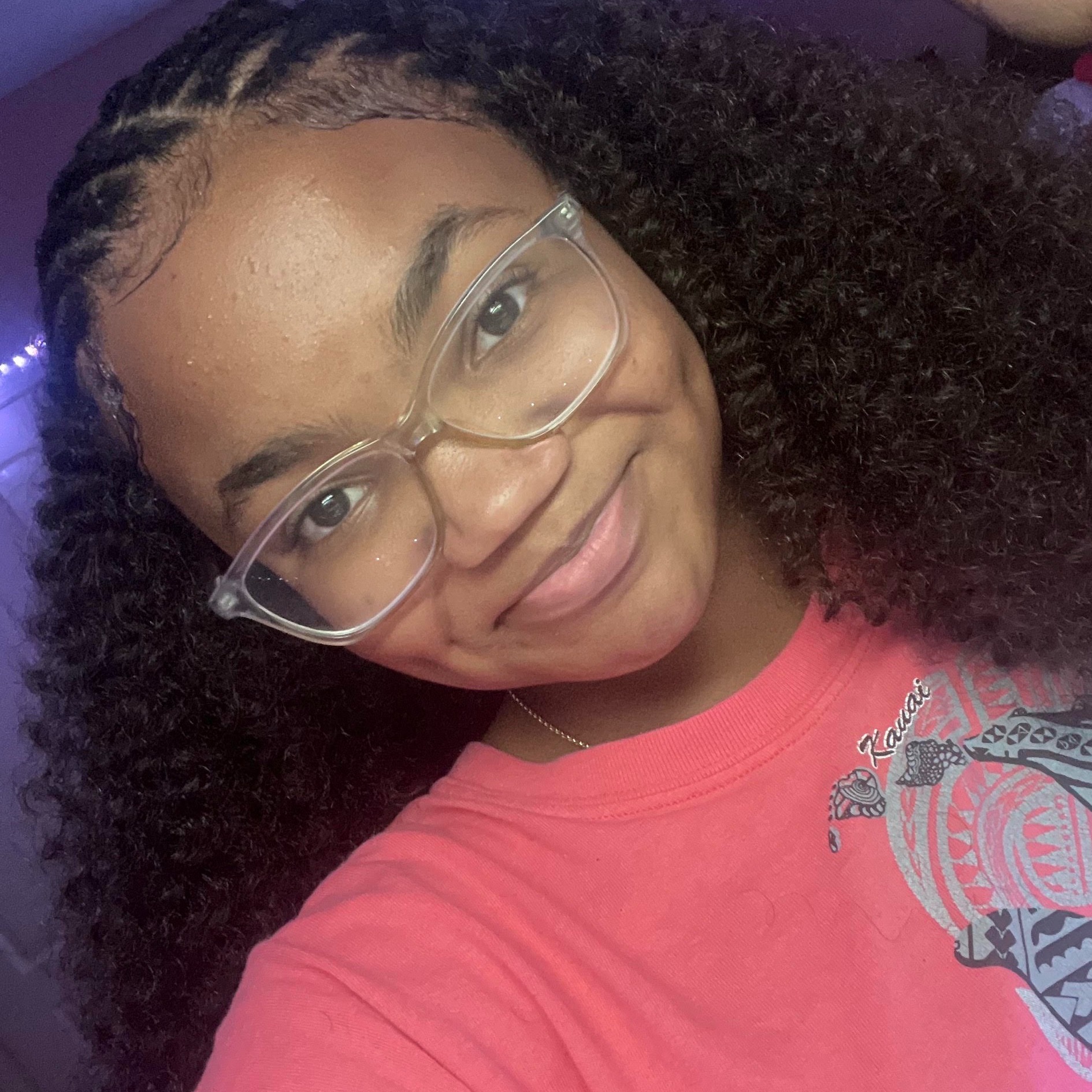 Person with curly hair, wearing glasses and a pink shirt, smiling. Background is dark.