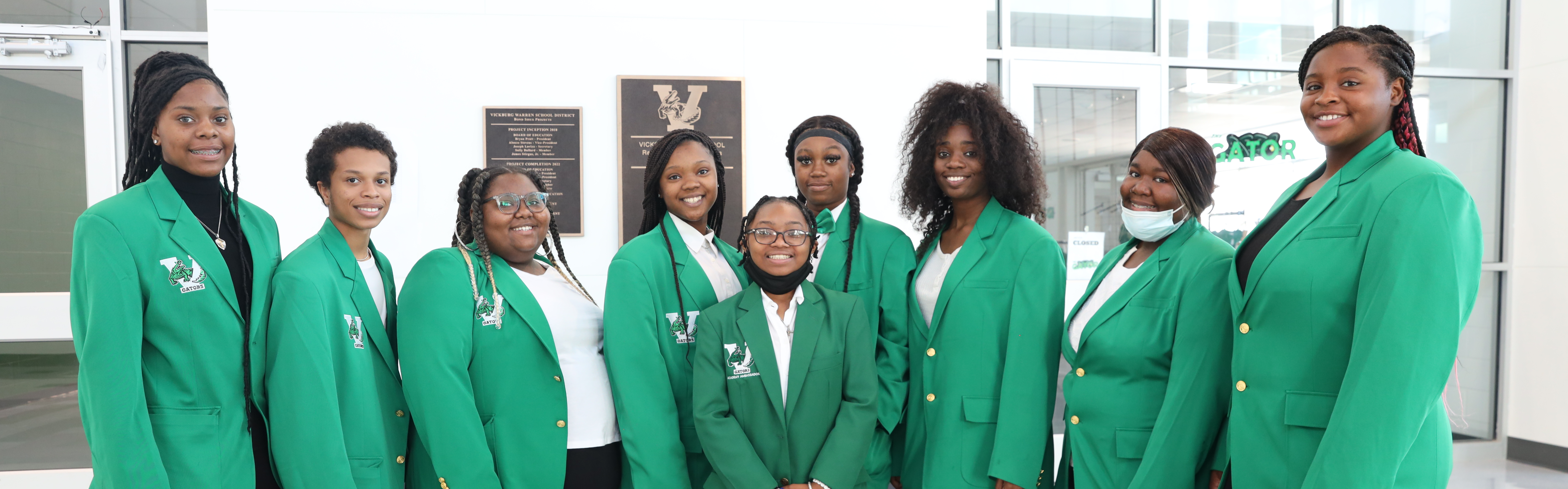 Nine high school students are posing for a photo while wearing green blazers and black bottoms.