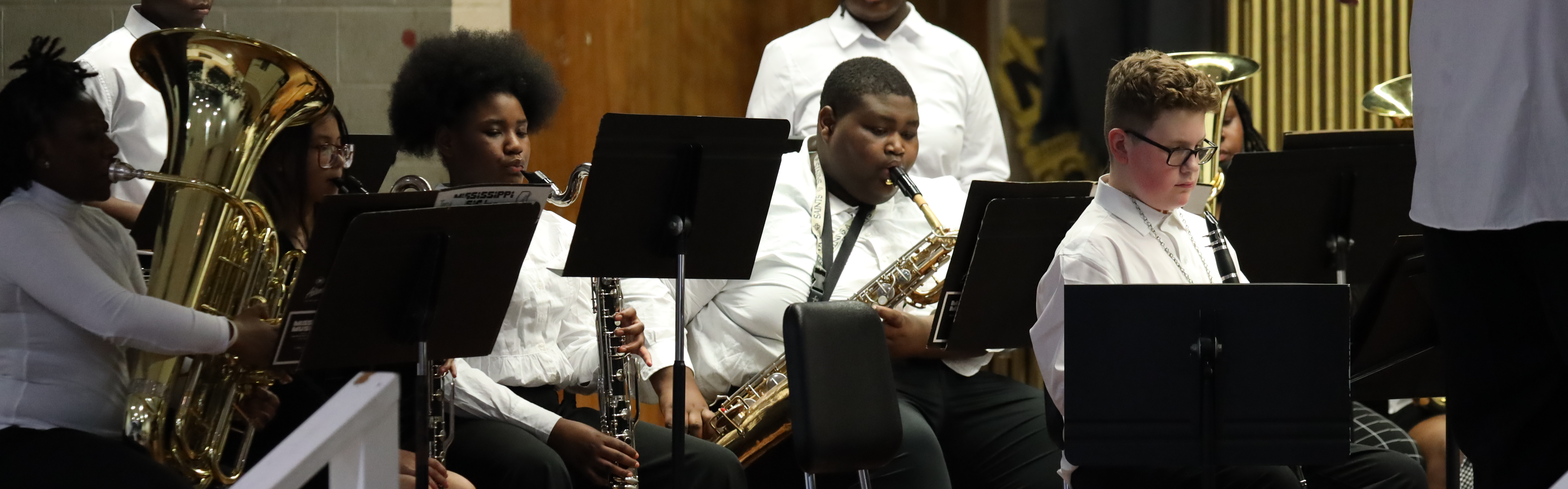 Four students are playing musical instruments in a concert ensemble. Two female students are playing bass clarinets, while the male student seated next to them is playing a baritone saxophone. Another male student is holding his clarinet.