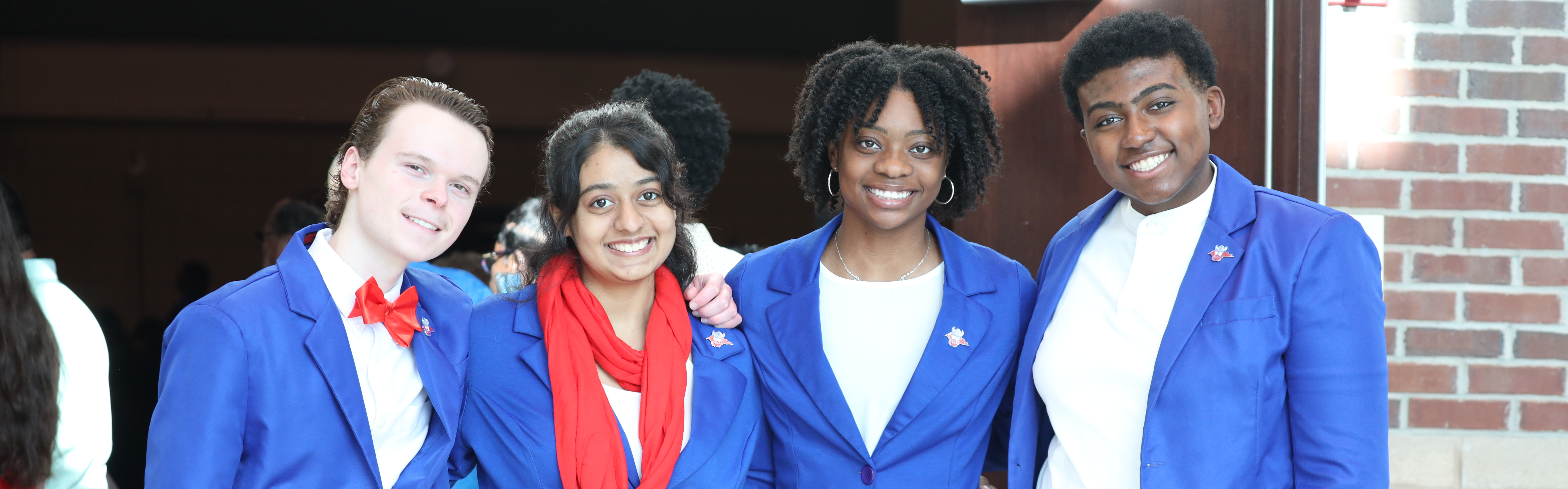 Two male and two female high school students pose for a group photo while wearing royal blue blazers and standing in a doorway.