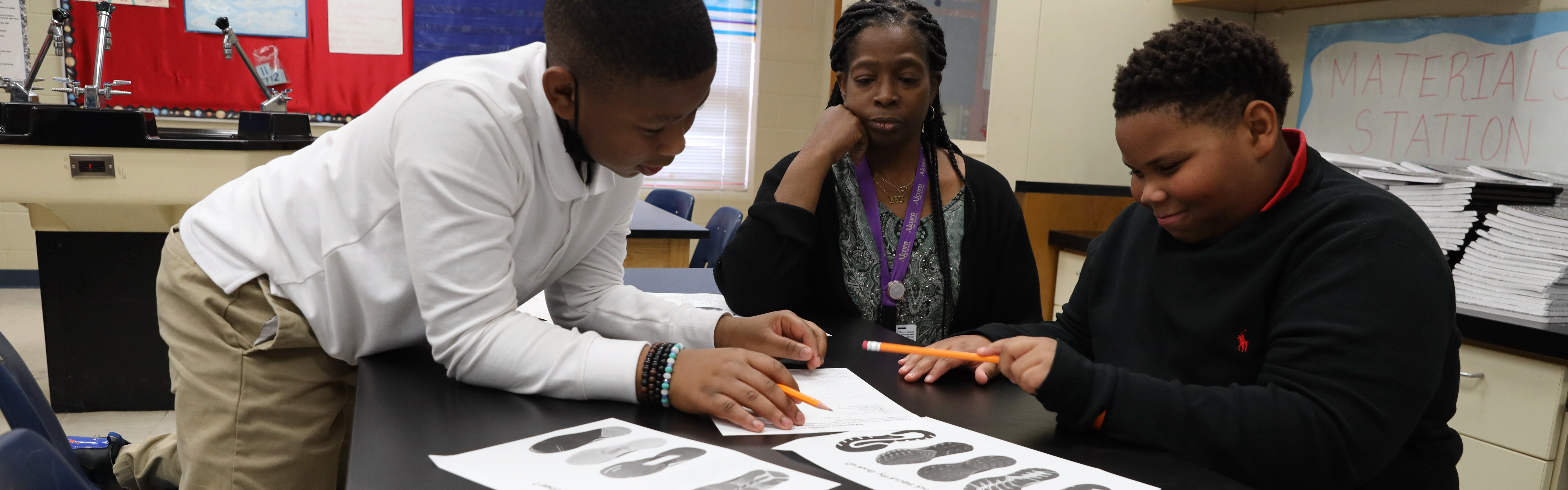 two african-american students work on a project on paper as their teacher observes. 