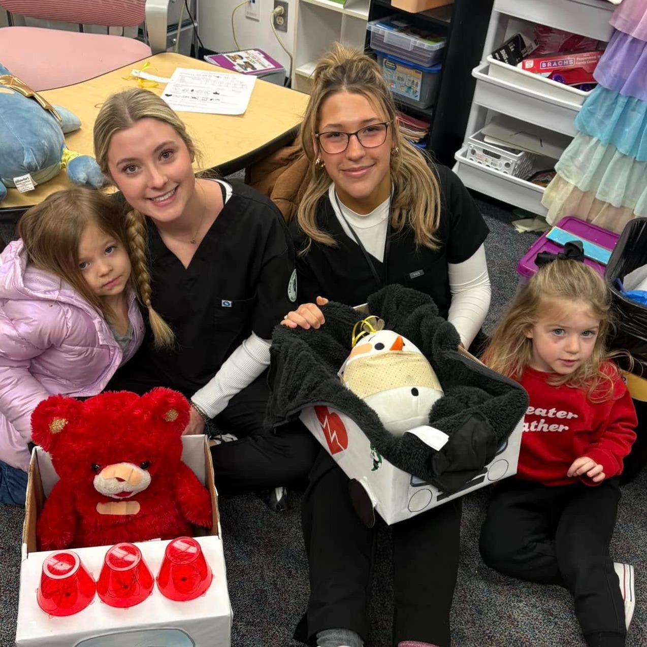 two high school girls wearing black scrubs with two young girls and teddy bears