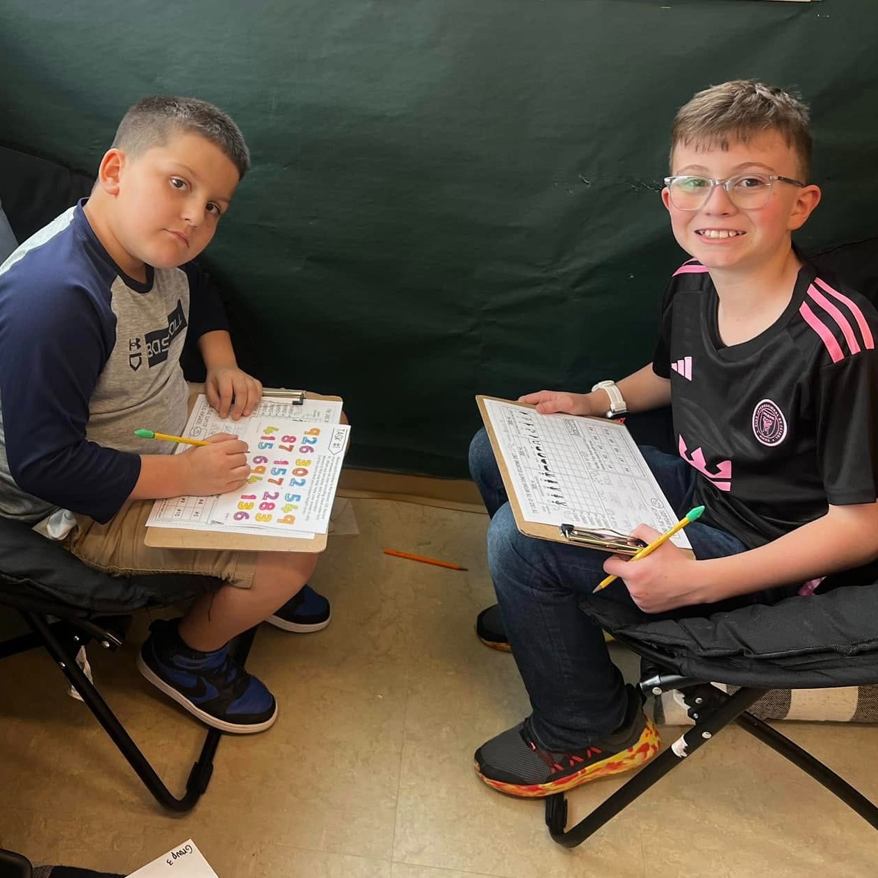 two boys sitting in chairs across from each other with clip boards