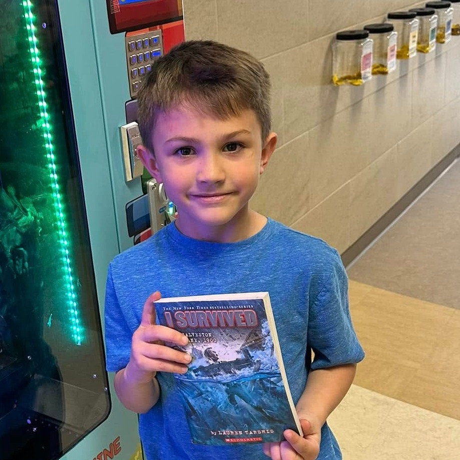 boy in blue shirt holding book next to a book vending machine