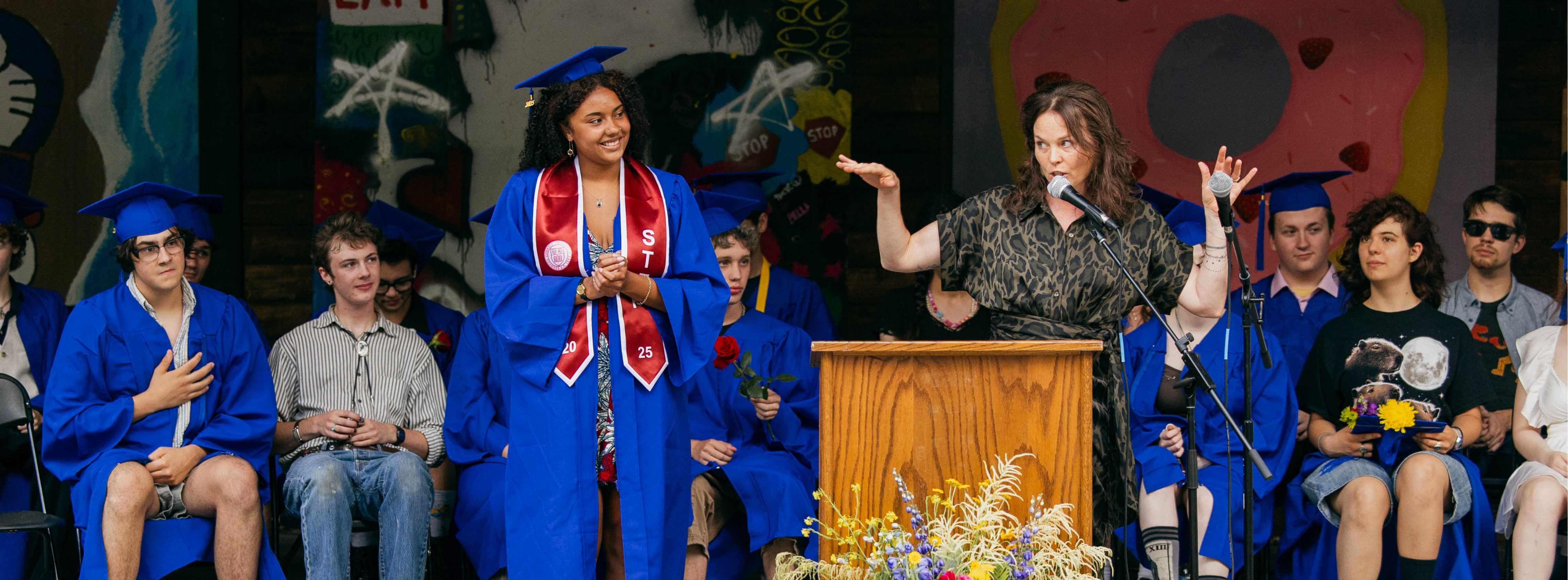 A woman speaks animatedly at a podium during a graduation ceremony, while a graduate in a blue cap and gown stands nearby, smiling and applauding. The stage is decorated with colorful artwork, including a large pink frosted donut painting and a mural featuring the word "EXIT." Seated behind them are other graduates in blue gowns and caps, along with casually dressed attendees. A vibrant floral arrangement is positioned in front of the wooden podium. The atmosphere is celebratory and expressive. 