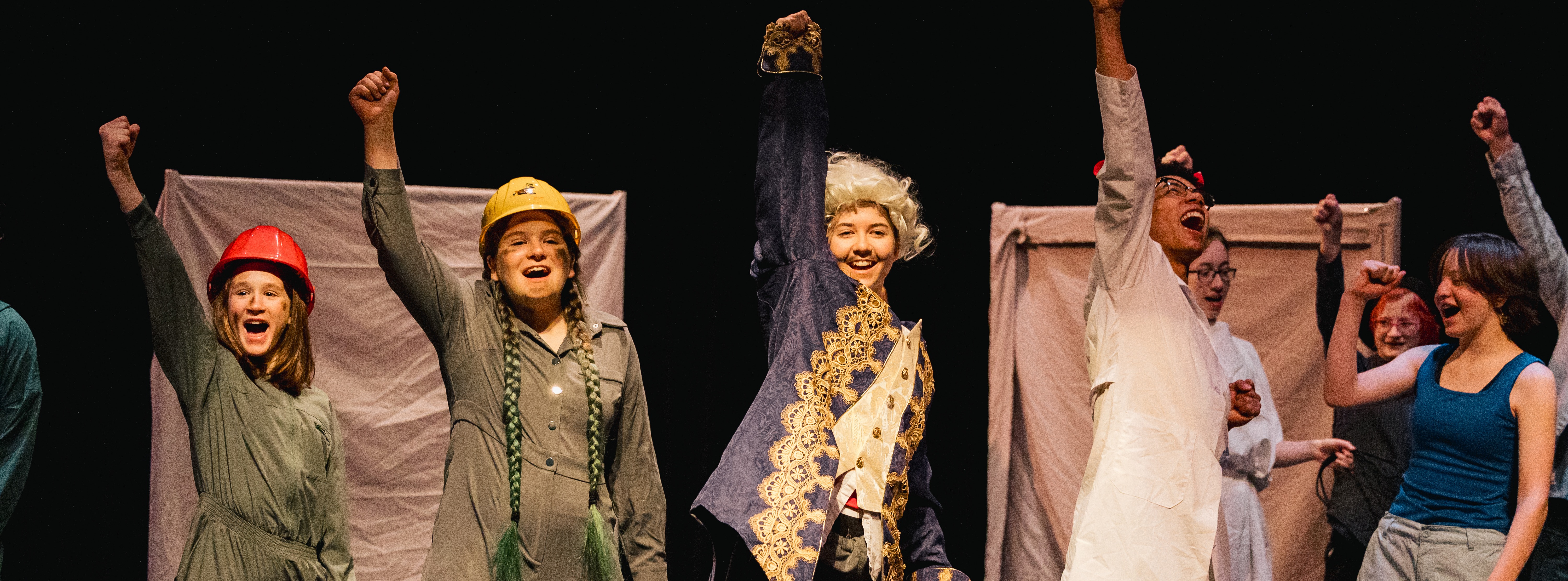 A group of actors performing on stage, raising their arms in the air, wearing various costumes.