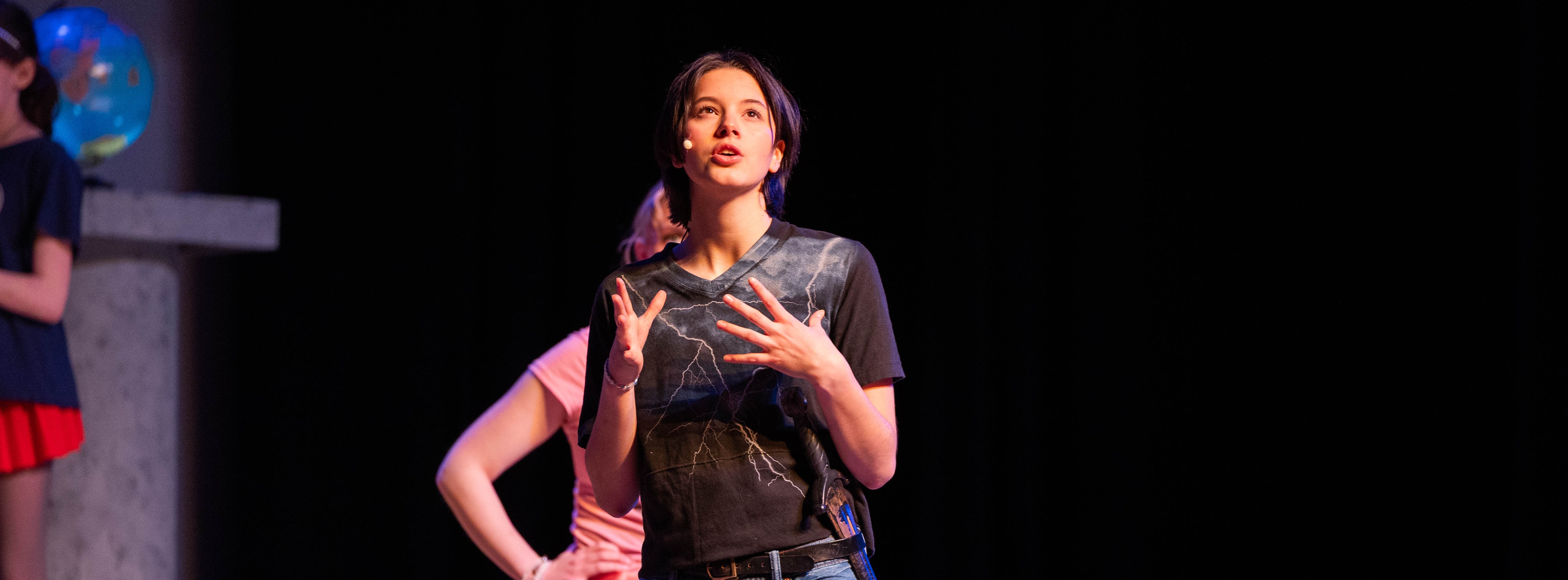 Two people on stage, one wearing a black t-shirt and jeans, the other in a pink shirt and red shorts.