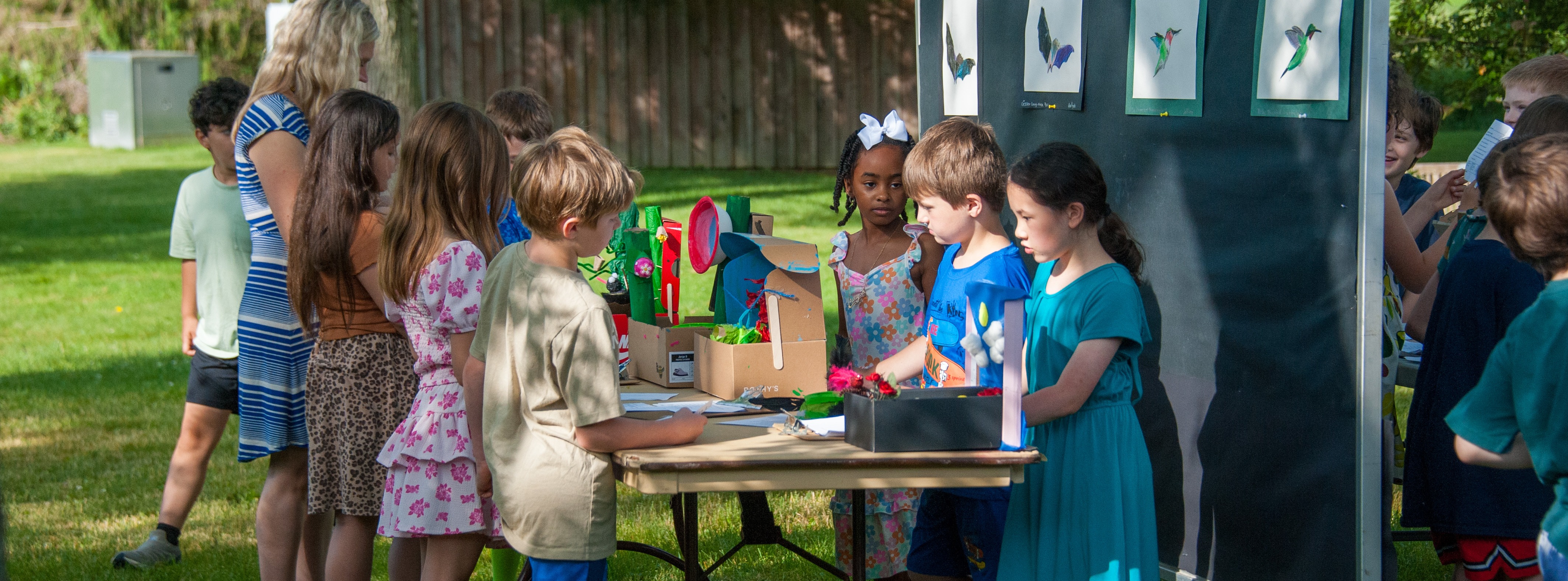 Kids gather around a table with educational materials in a grassy outdoor area. A backdrop displays pictures of insects.