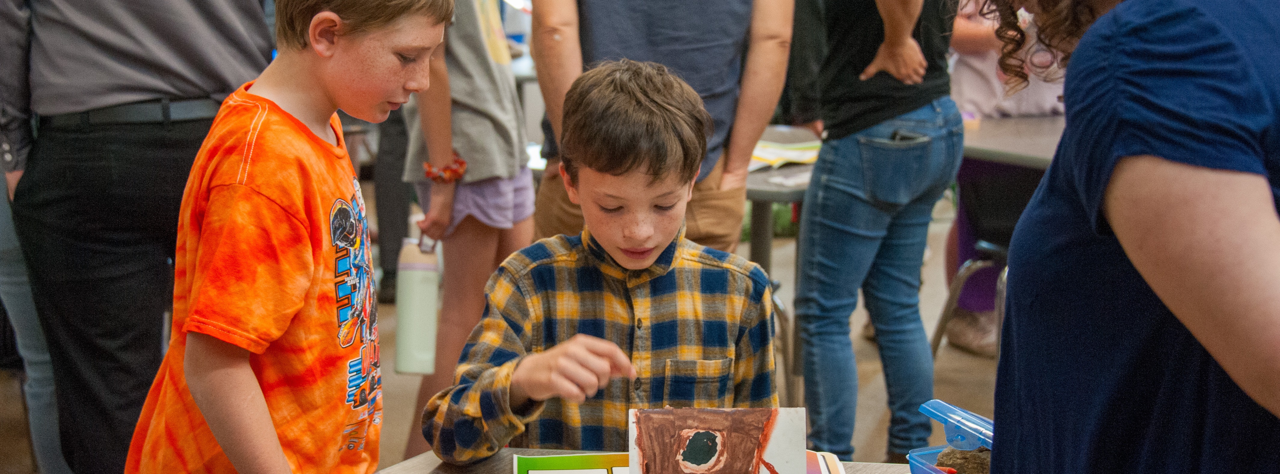 Three children stand around a table displaying a model. The child in orange looks at the model.