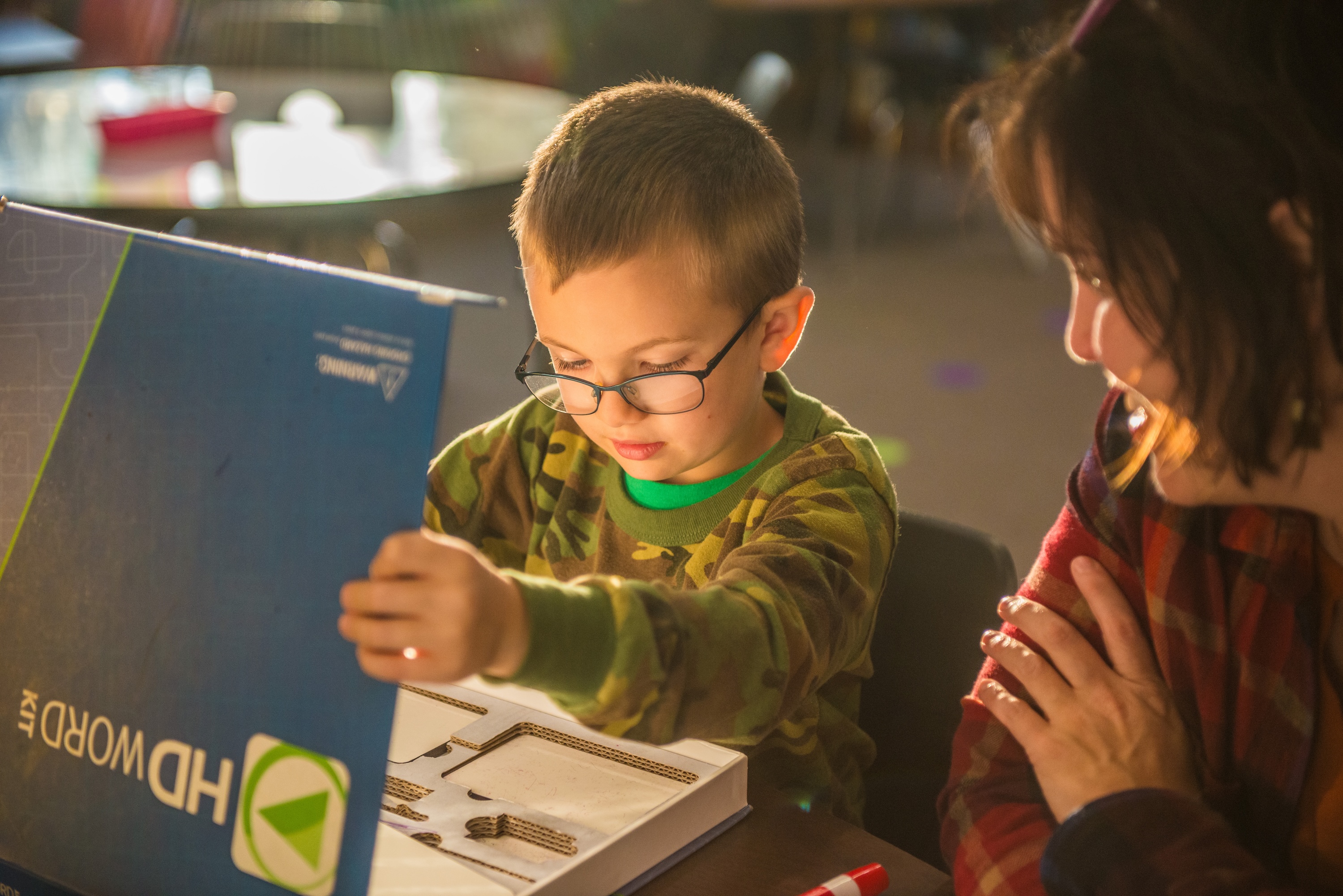 A boy in glasses examines a blue box while a woman watches, seated at a table.