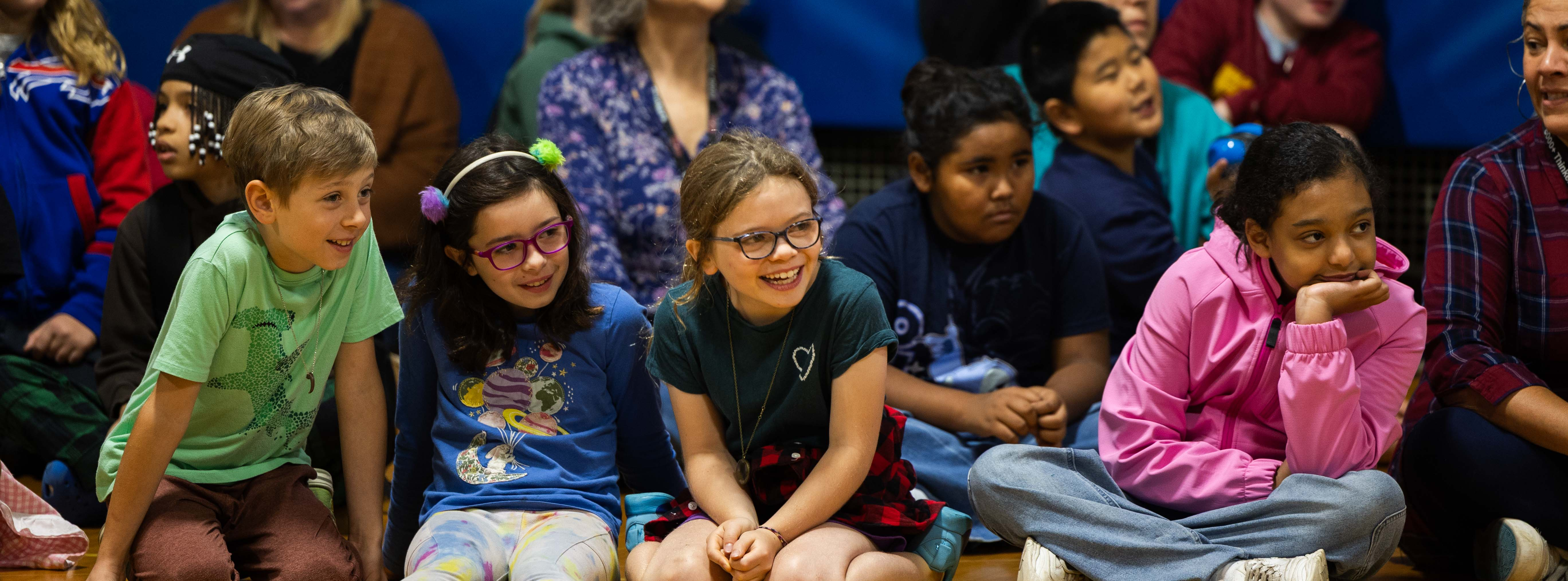 A group of students and several adults sitting on the gym floor, watching a performance, and laughing
