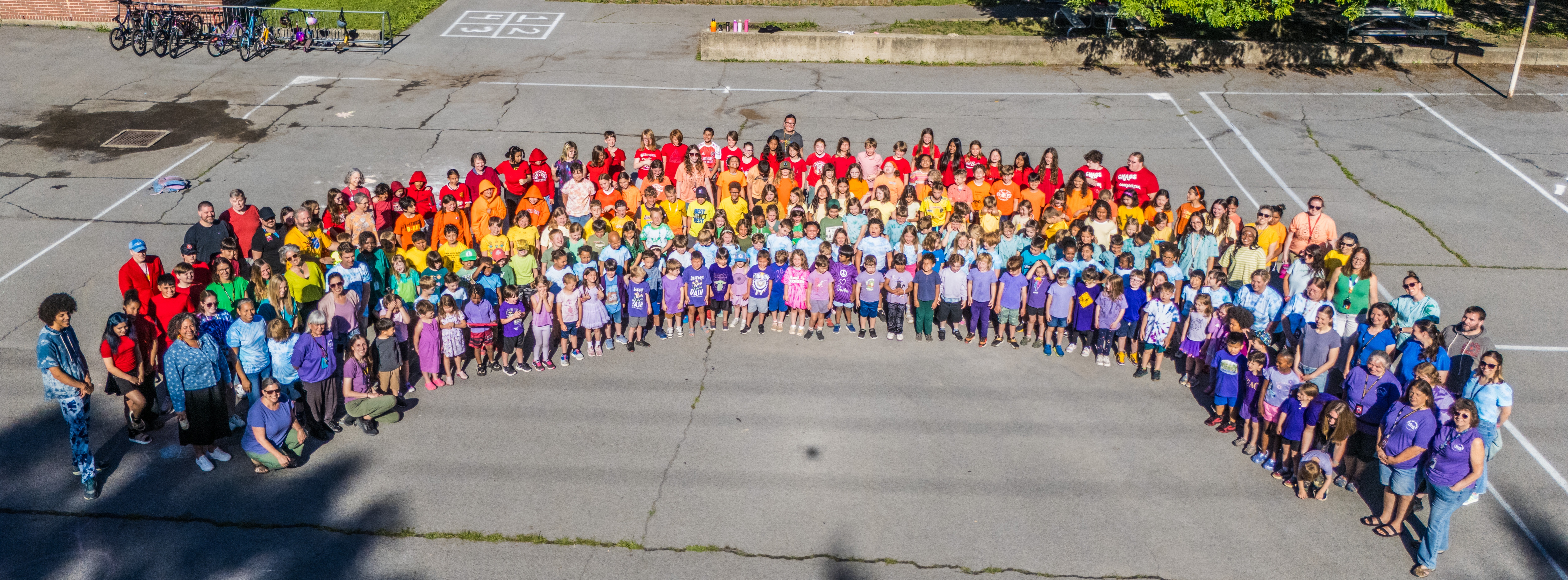 A large group of people wearing various colors form a rainbow shape in a parking lot.