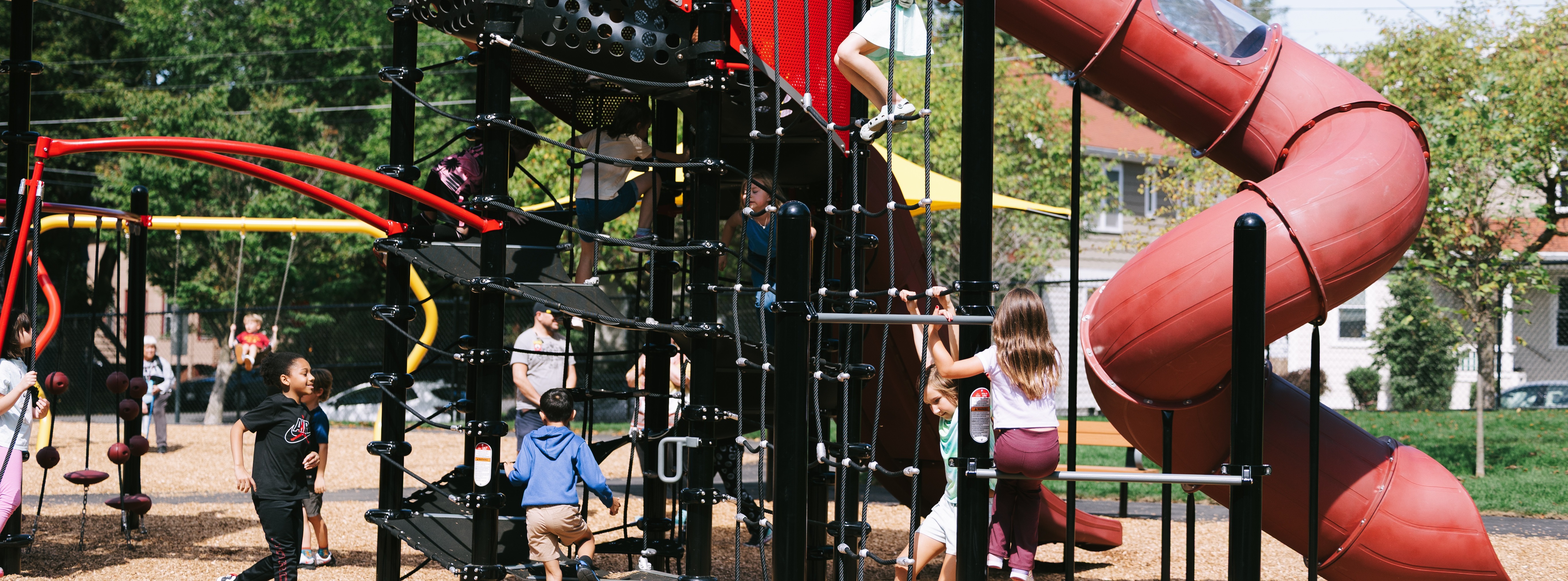 Children play on a large, red slide and climbing equipment at an outdoor playground under a blue sky.