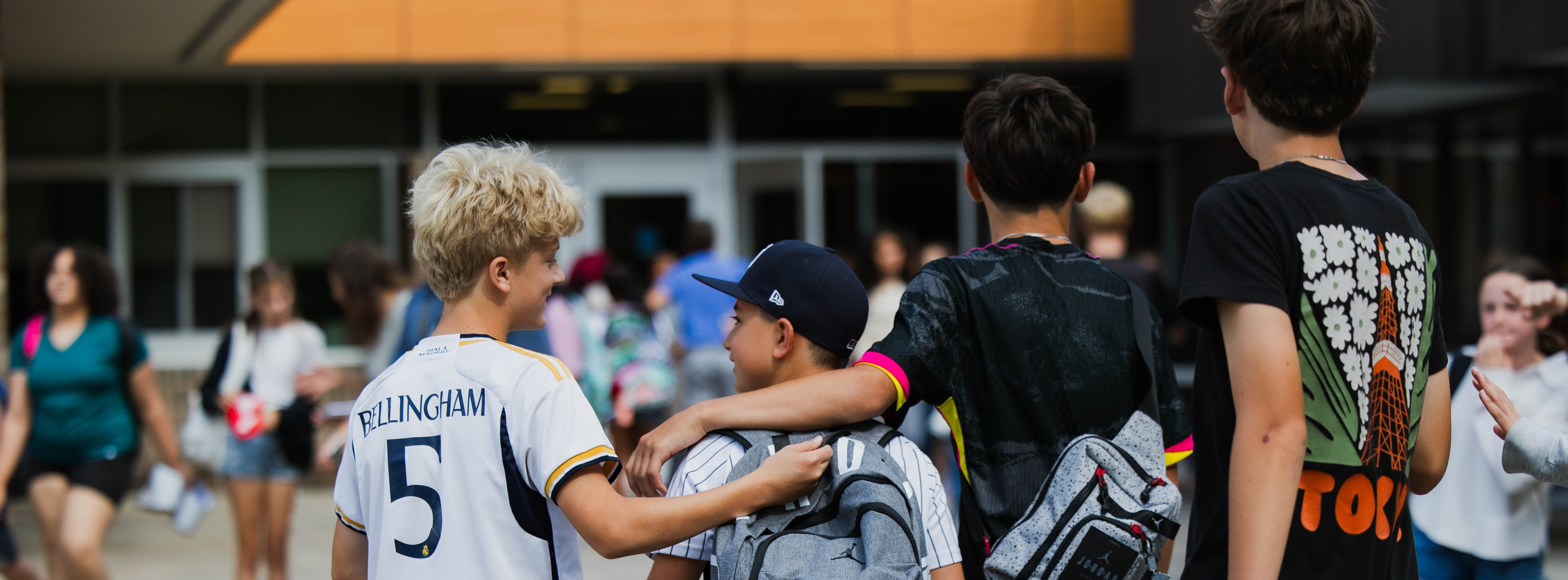 Four boys with backpacks walk together outside a school, with other students in the background