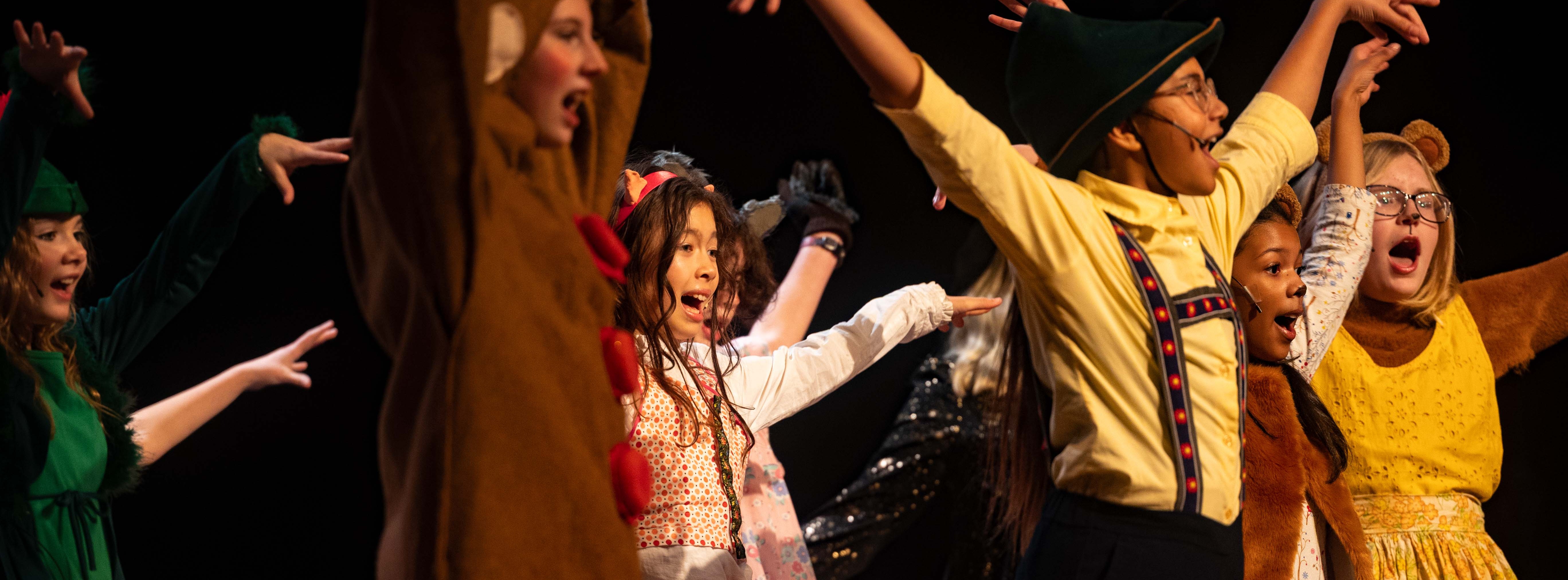 Children in costumes perform on a stage. Characters include a bear, witch, and others.
