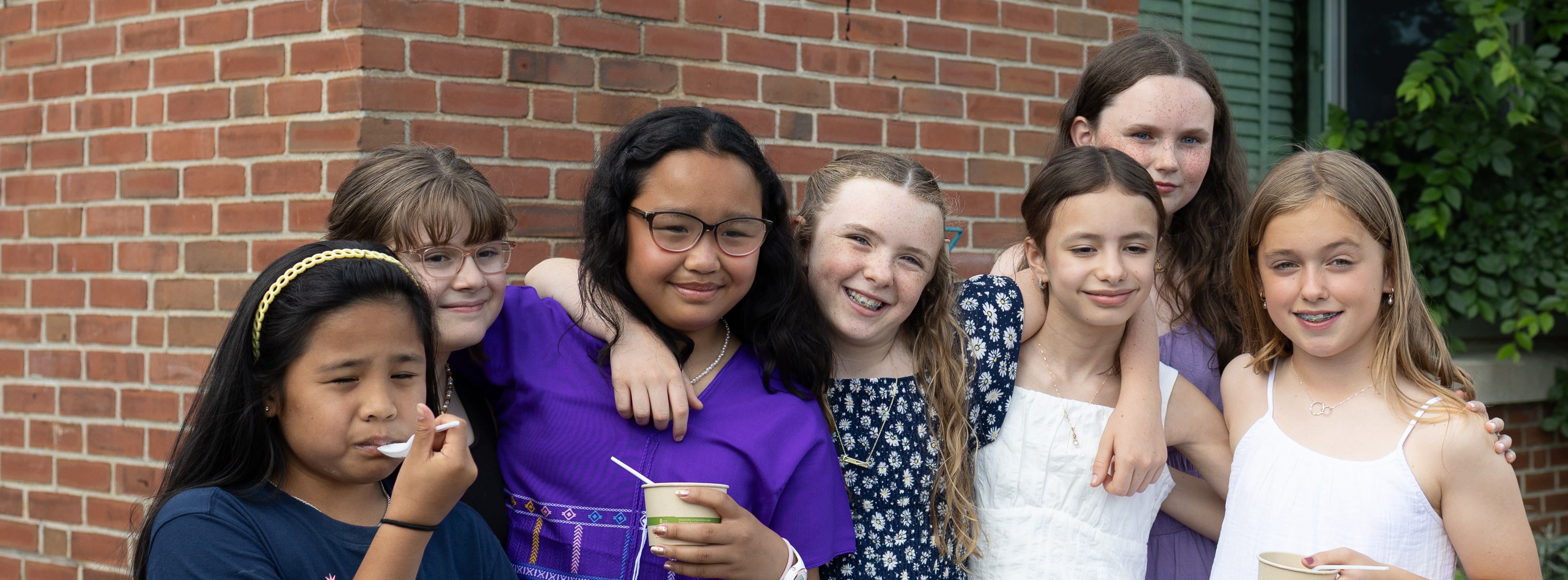A group of children posing for a photo, holding cups and straws, with brick wall in the background.