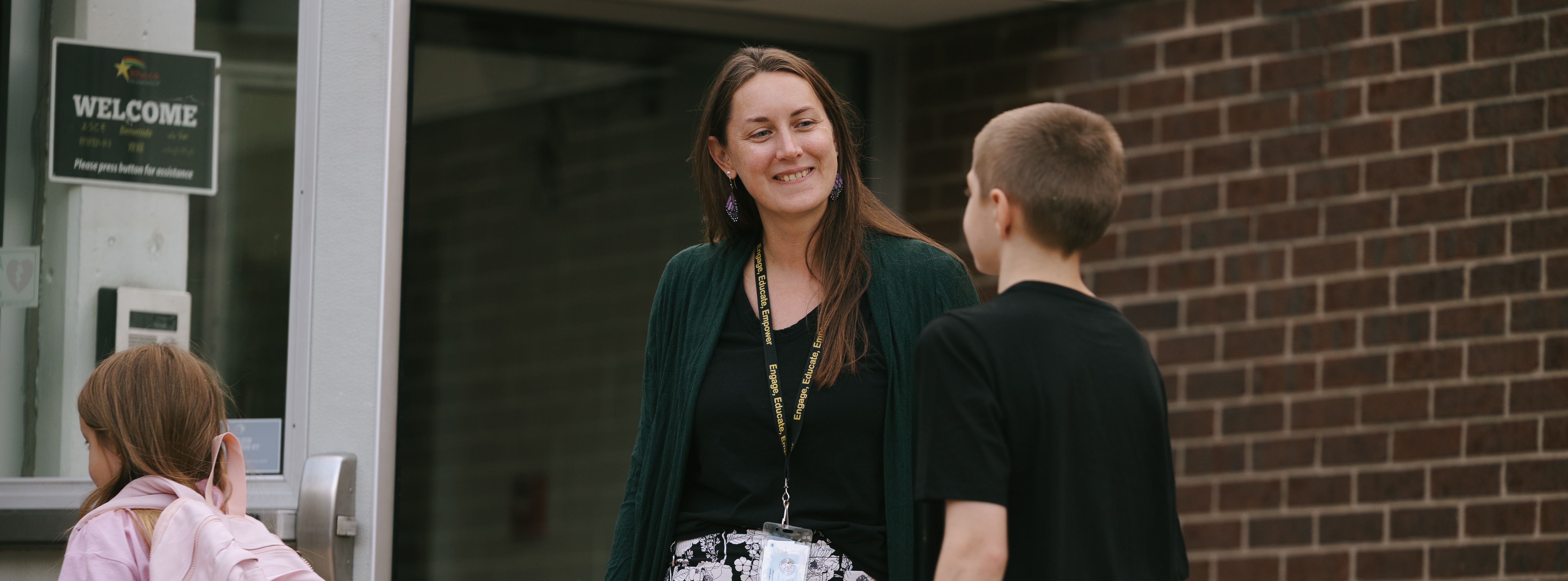 A woman with a lanyard speaks to a boy and girl outside a building with a "Welcome" sign.