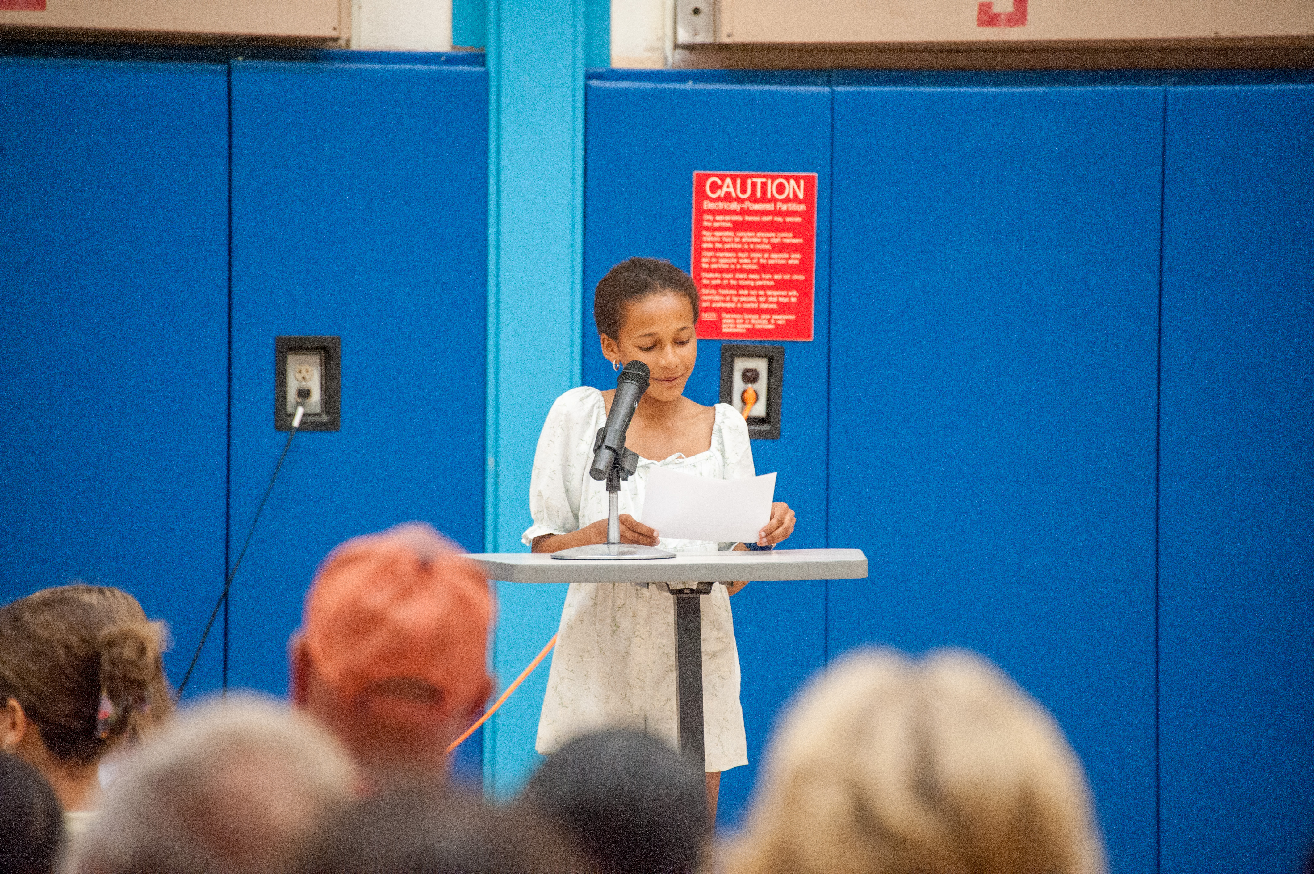 A woman reads from paper, standing at a podium with a microphone in front of a blue wall.