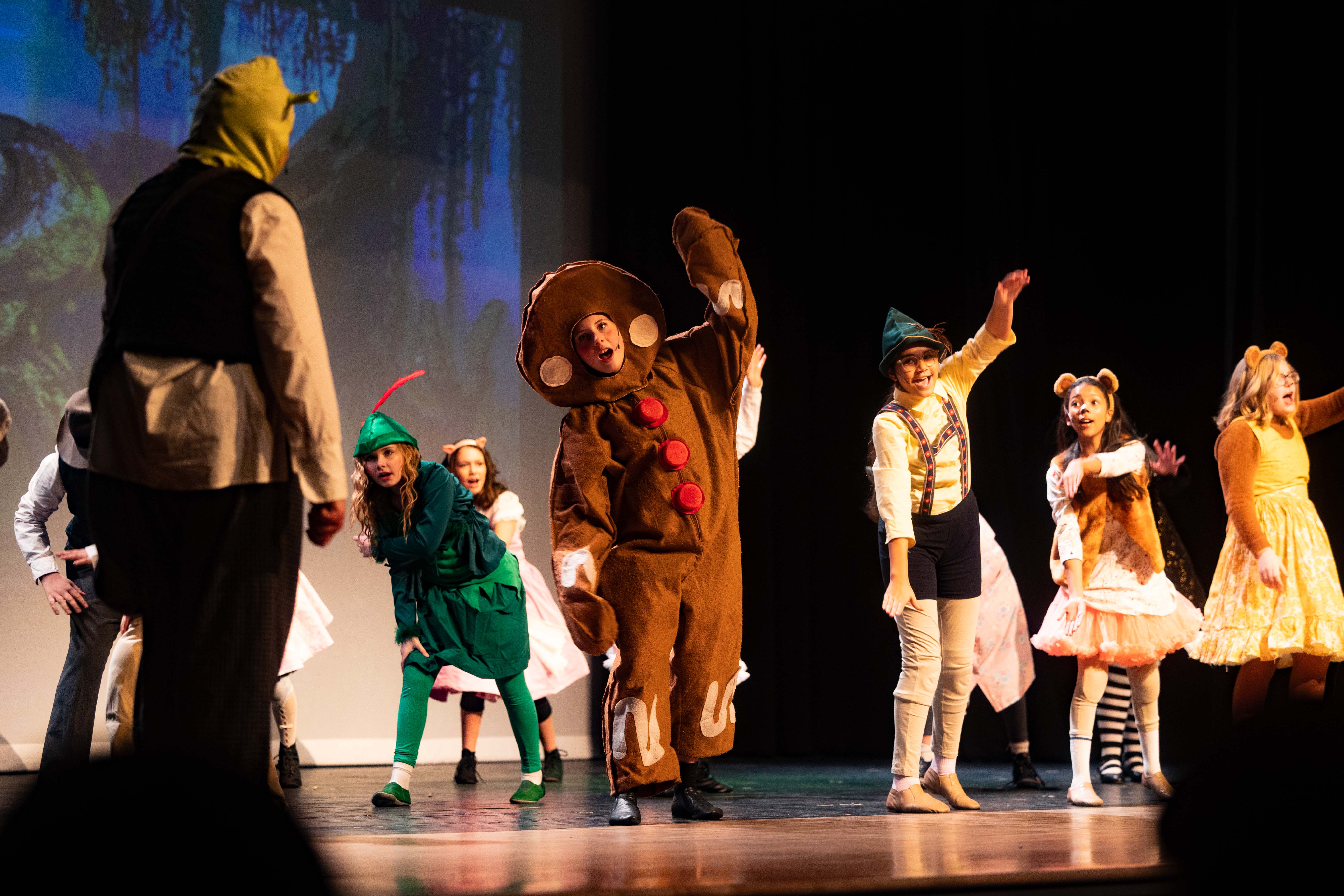 A group of children perform a play on stage. They wear costumes, and one person is in a gingerbread costume.