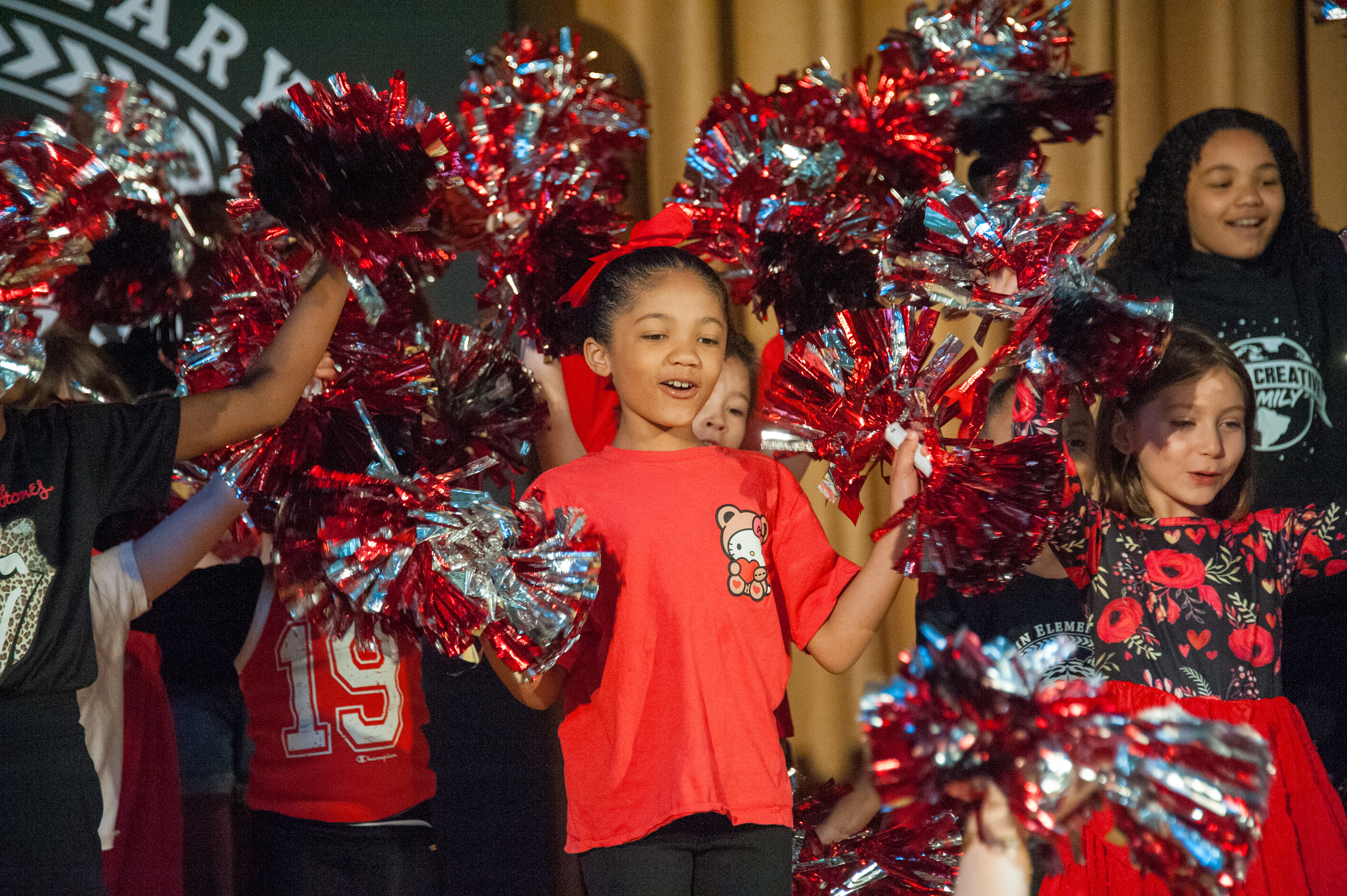 Group of children wearing red shirts and black pants, holding red and silver pom-poms, standing and performing.