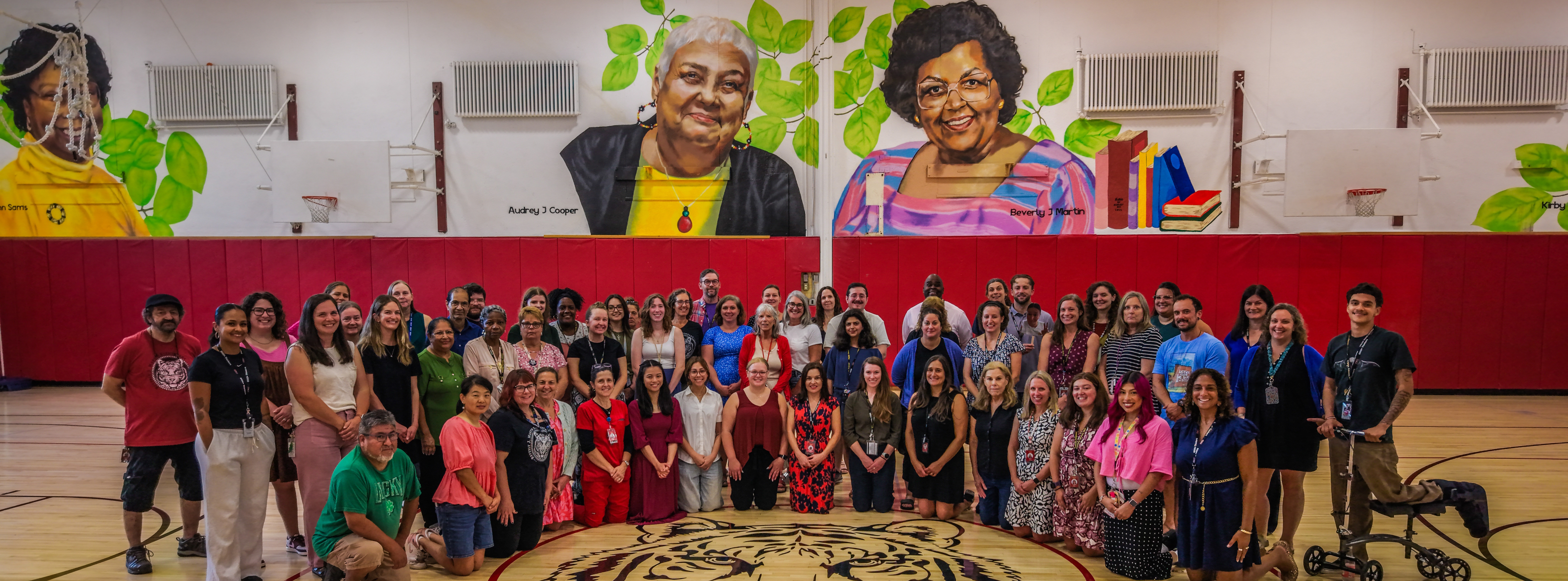 A group of people stand around a circular tiger design on a basketball court. Two large portraits adorn the background.