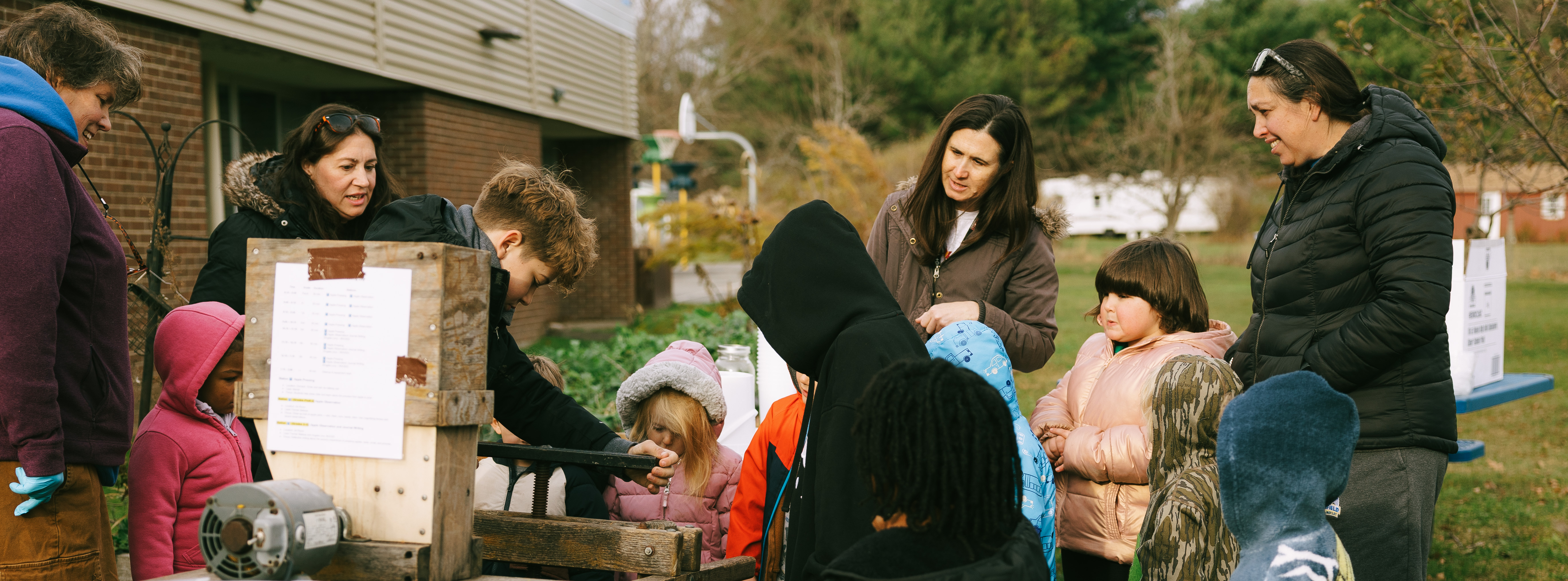 A group of children and adults gather outdoors around an old-fashioned wooden apple cider press. The kids, bundled in colorful winter coats and hats, watch as apples are pressed. Bowls of apples sit on a nearby blue-covered table, and the activity takes place beside a brick school building with trees and greenery in the background.