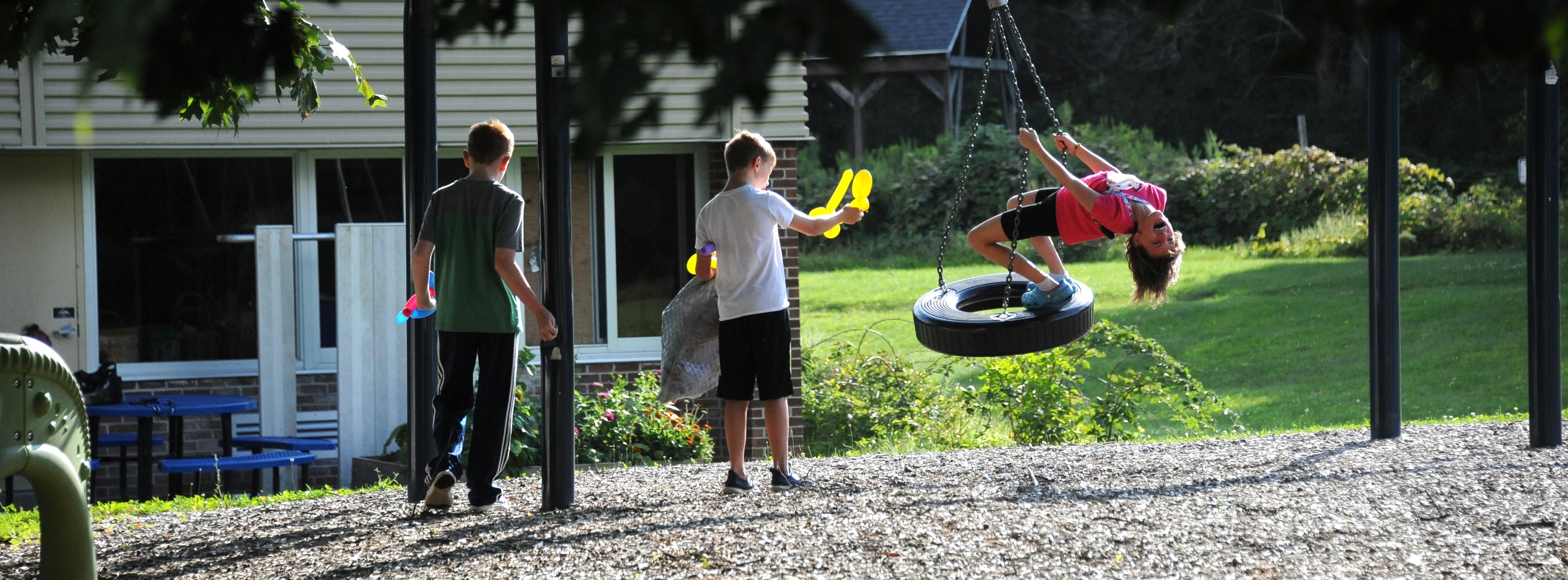A playground with a tire swing and gravel ground. One child swings while others stand nearby.