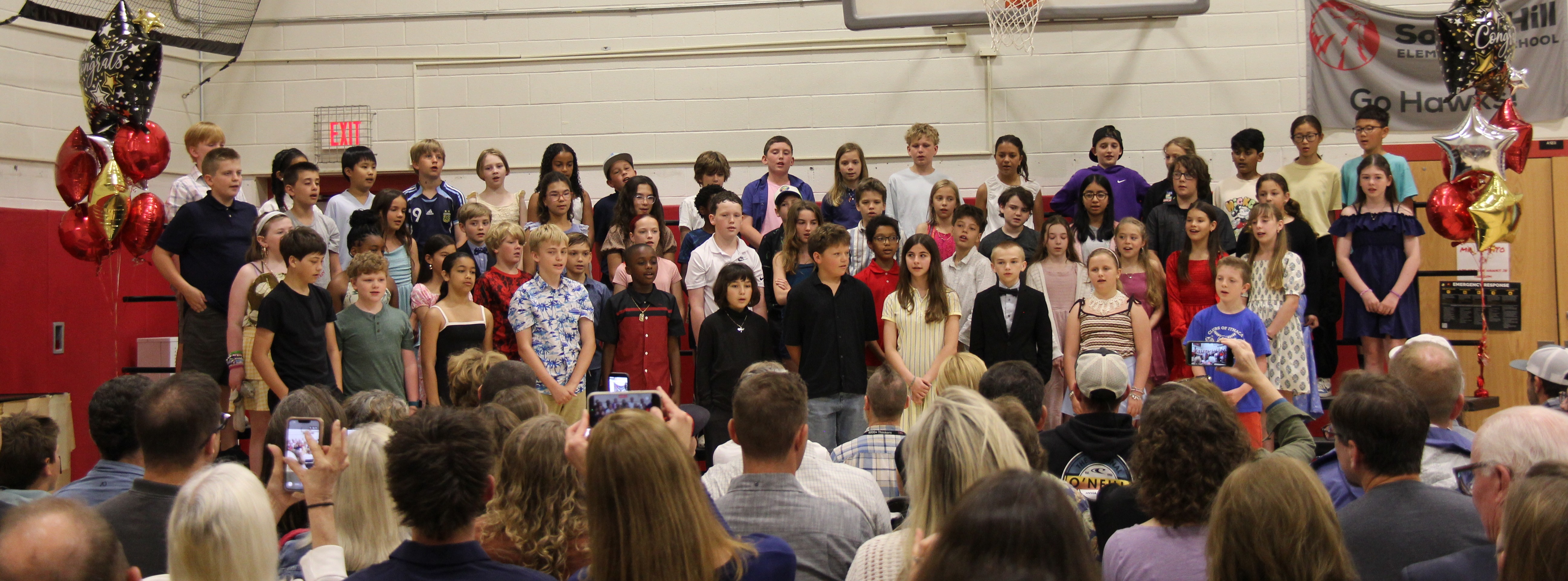 A group of students standing on a stage in a gymnasium with a basketball hoop. Audience seated in front.