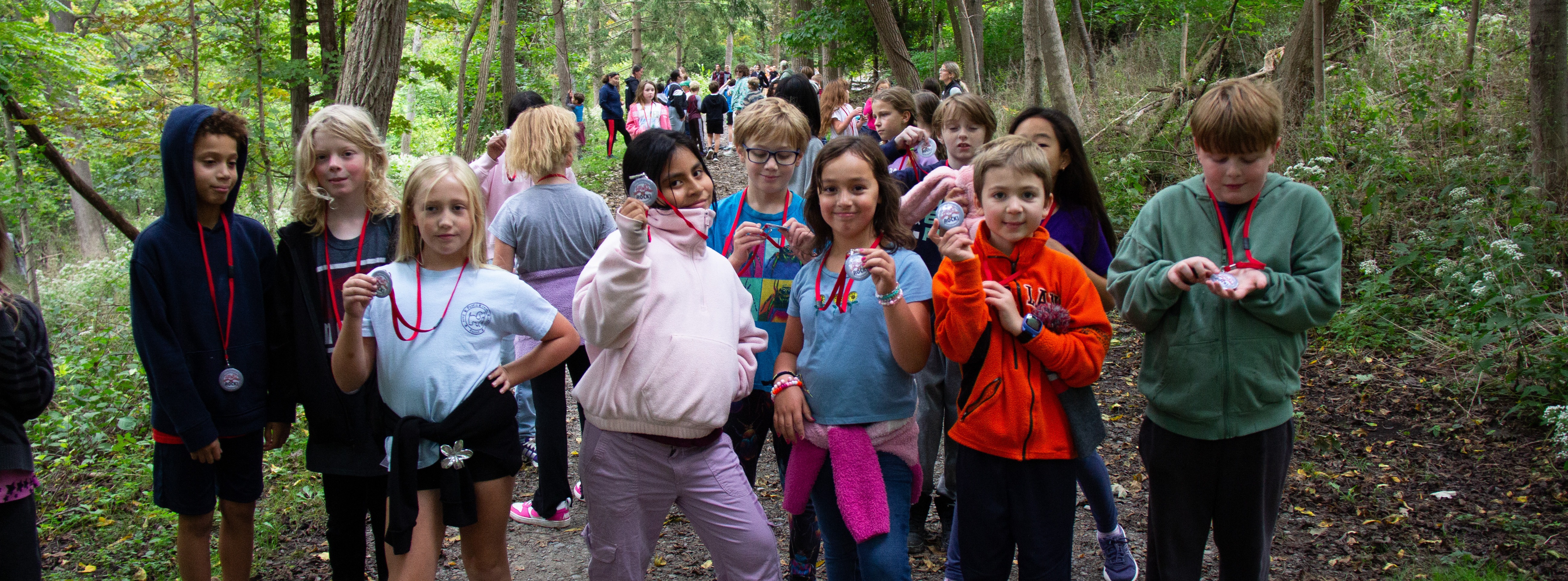 A group of children stand in a forest path, wearing medals. Trees and greenery surround them.