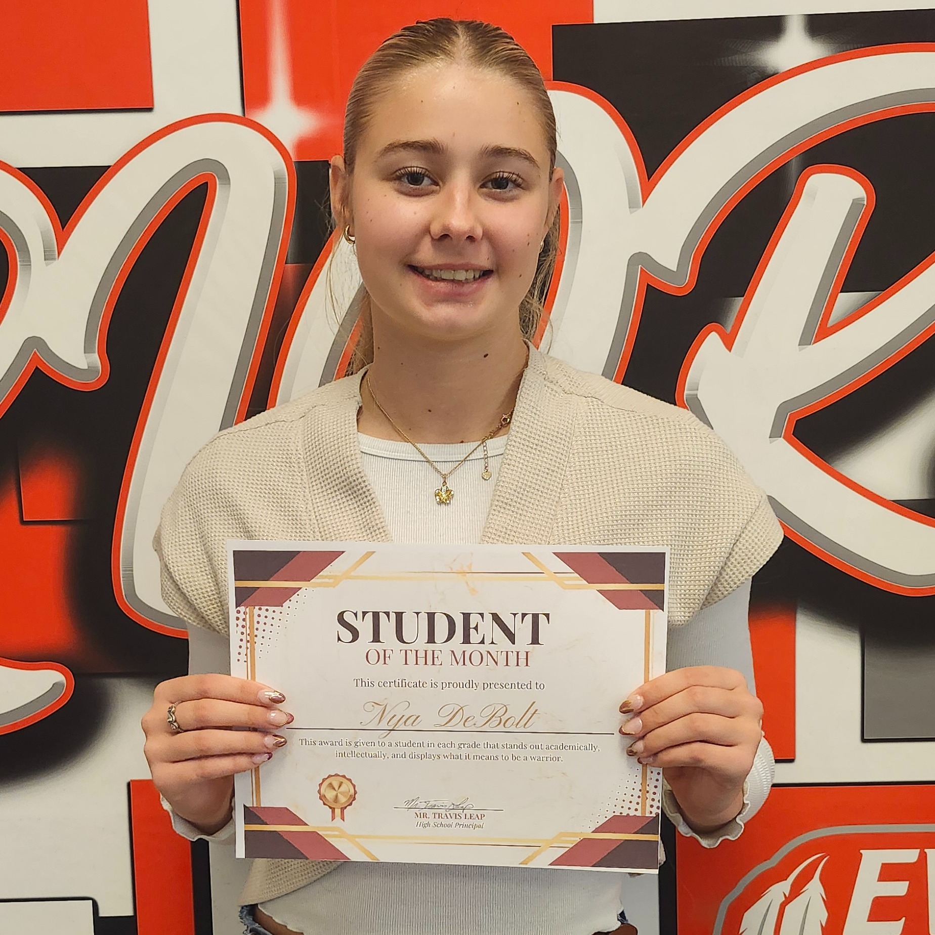 A student stands before a wall with Everett Warriors logo, holding a certificate with "STUDENT" in bold letters.