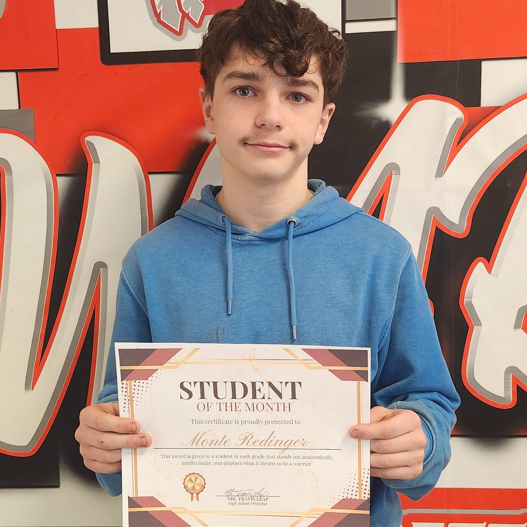 A young boy in a blue hoodie holding a framed certificate, standing in front of a wall with school logos.
