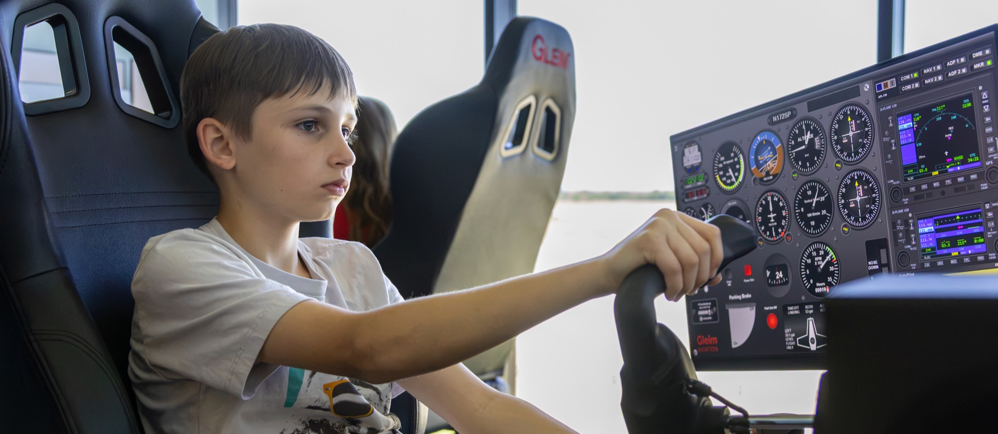 A student sits focused in a pilot seat, using a full flight simulator yoke and digital instrument panel display.