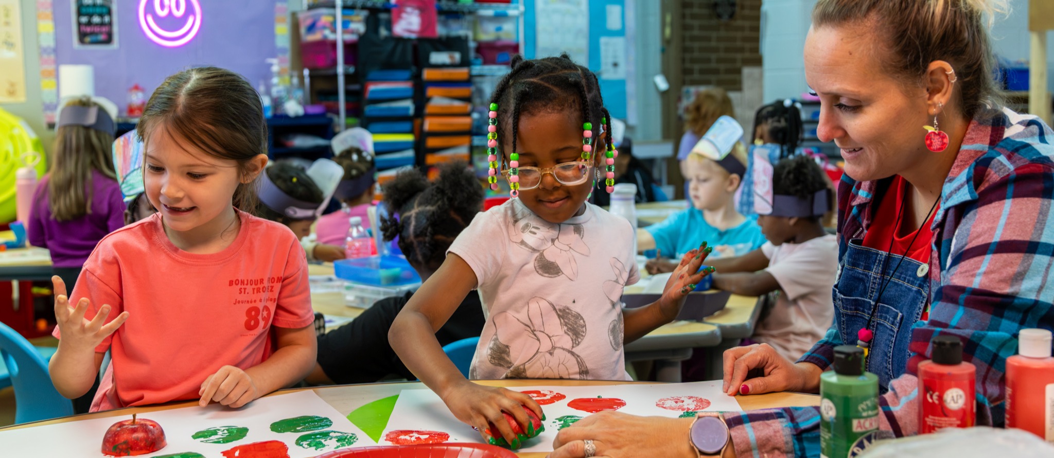 Two students use cut apples and paint to stamp shapes onto a piece of paper as their teacher encourages them.