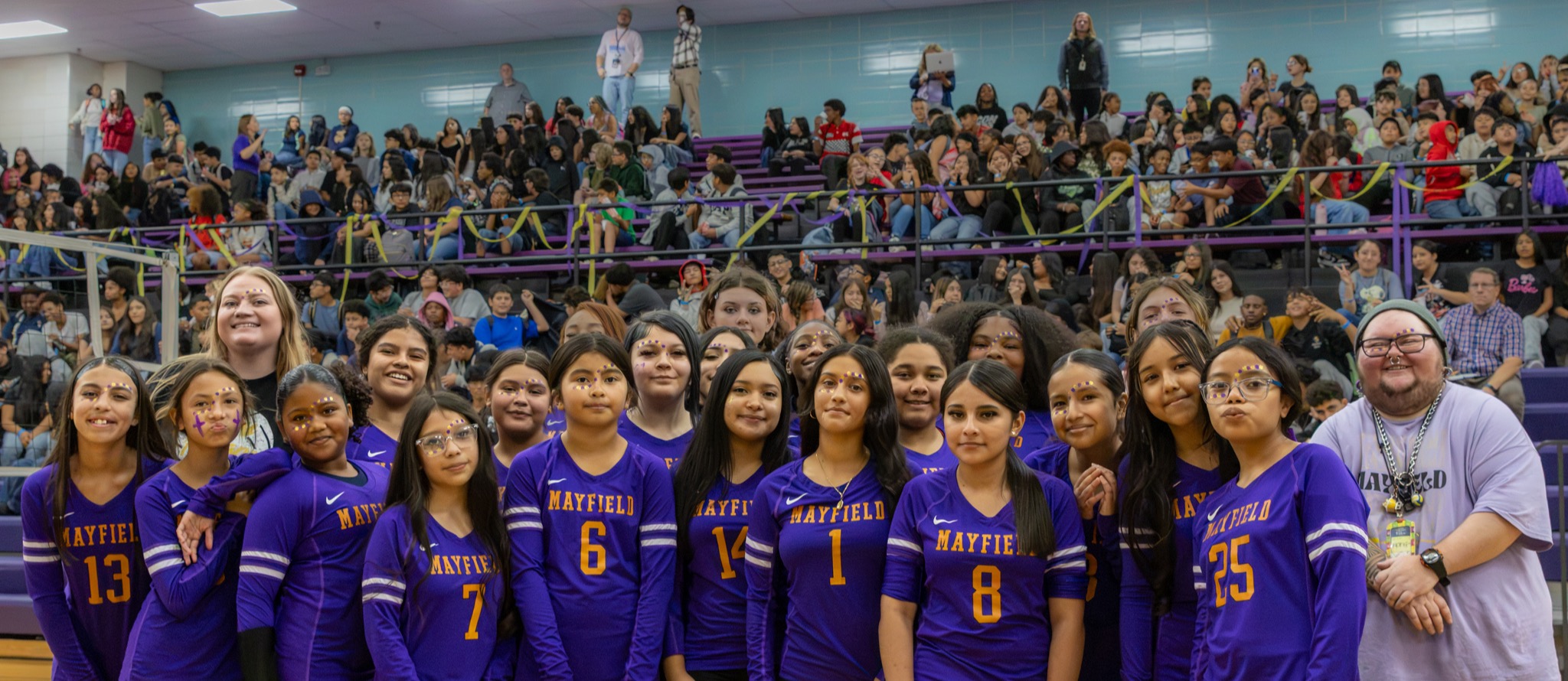 Mayfield Middle School volleyball players and staff pose together in purple and gold in front of a packed gymnasium. Students wearing purple and yellow face decorations stand shoulder to shoulder, with classmates filling the bleachers behind them.
