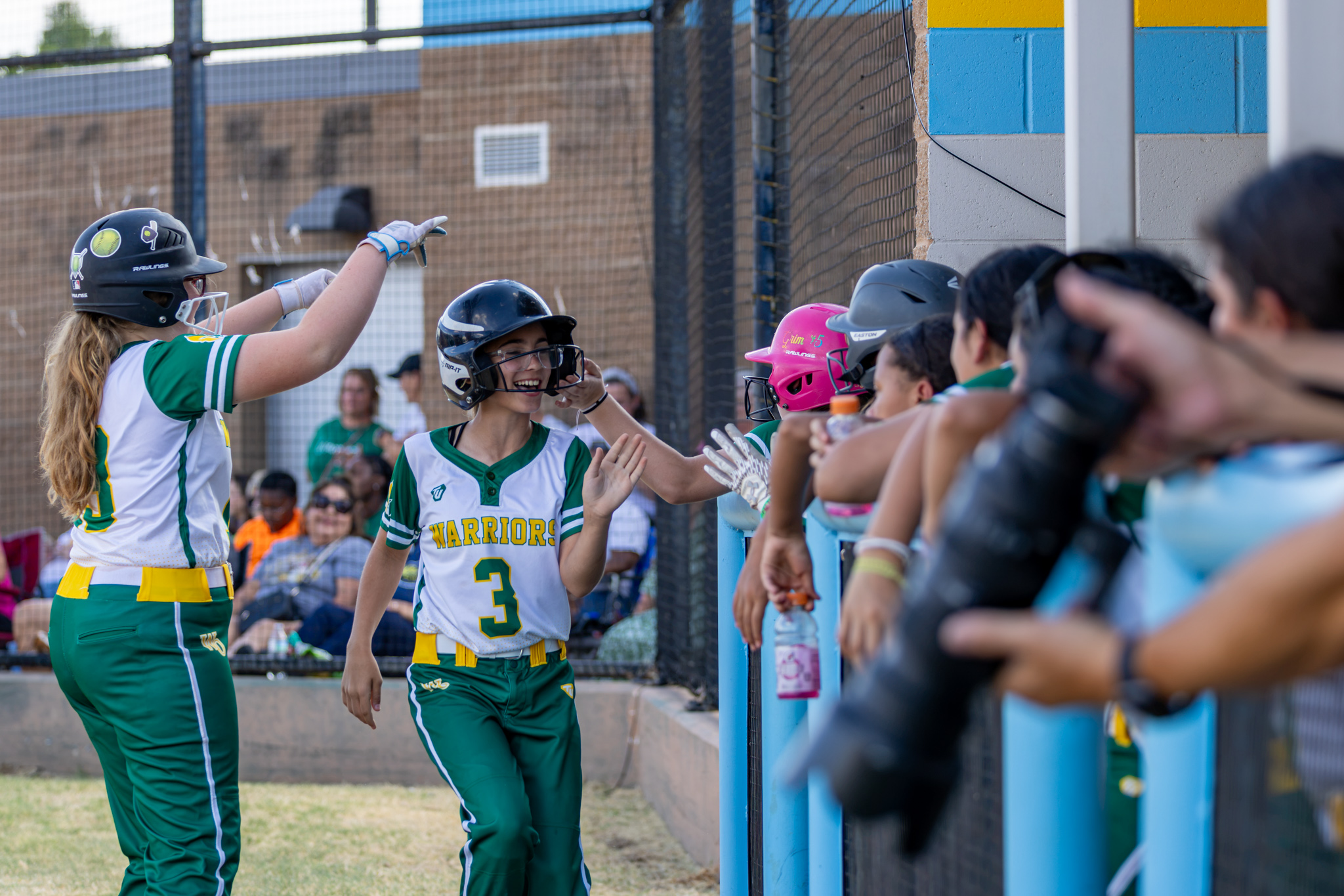 A WOMS softball player runs back to the dugout after scoring a run, with teammates cheering her on.