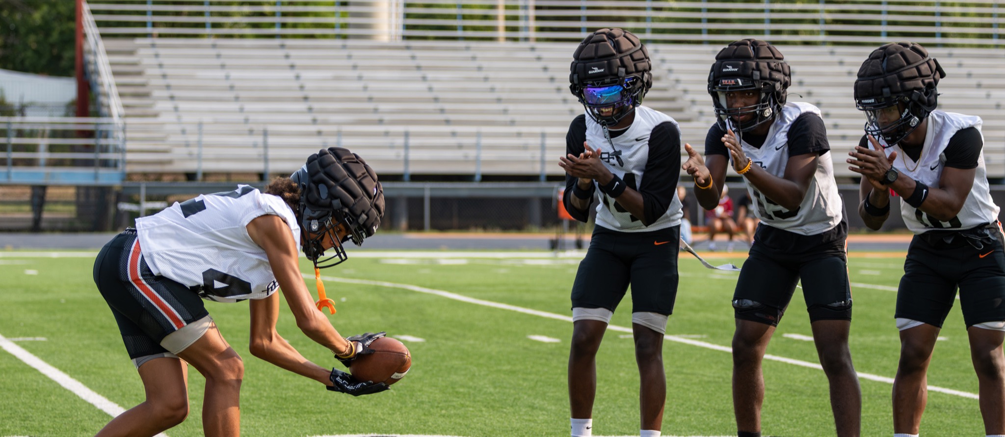 Putnam City High football players practice a snap drill as three teammates clap and wait in position on the field.
