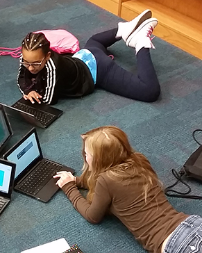 Two teens on the floor using laptops.