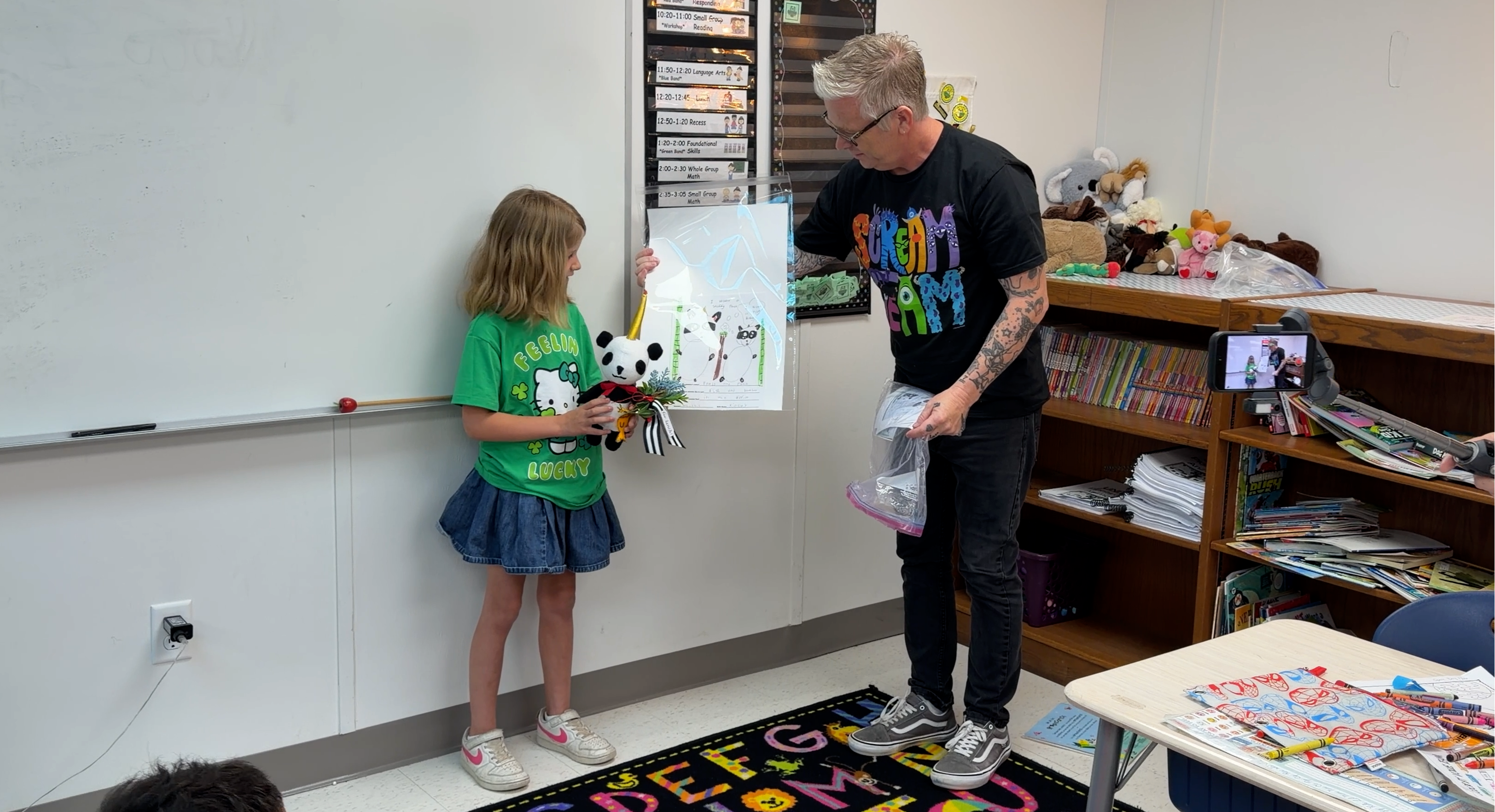 A man presents a drawing and a panda plushie to a smiling young girl in a colorful classroom.