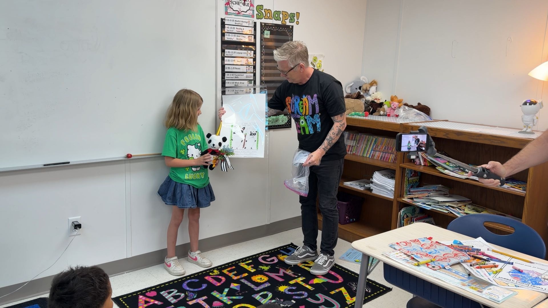 A man presents a drawing and a panda plushie to a smiling young girl in a colorful classroom.