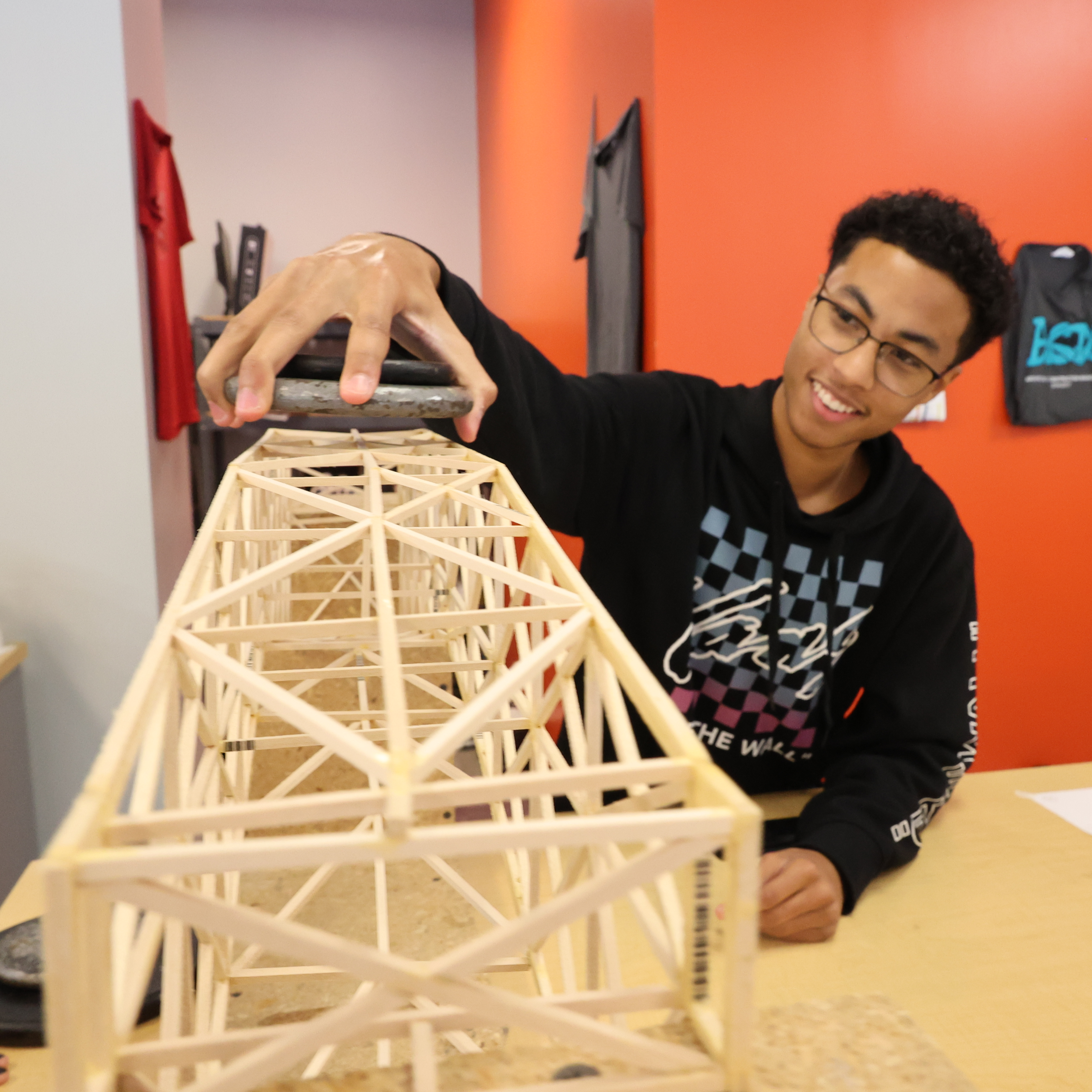 Black, male student holds a small circular weight over a bridge constructed by small wooden sticks.