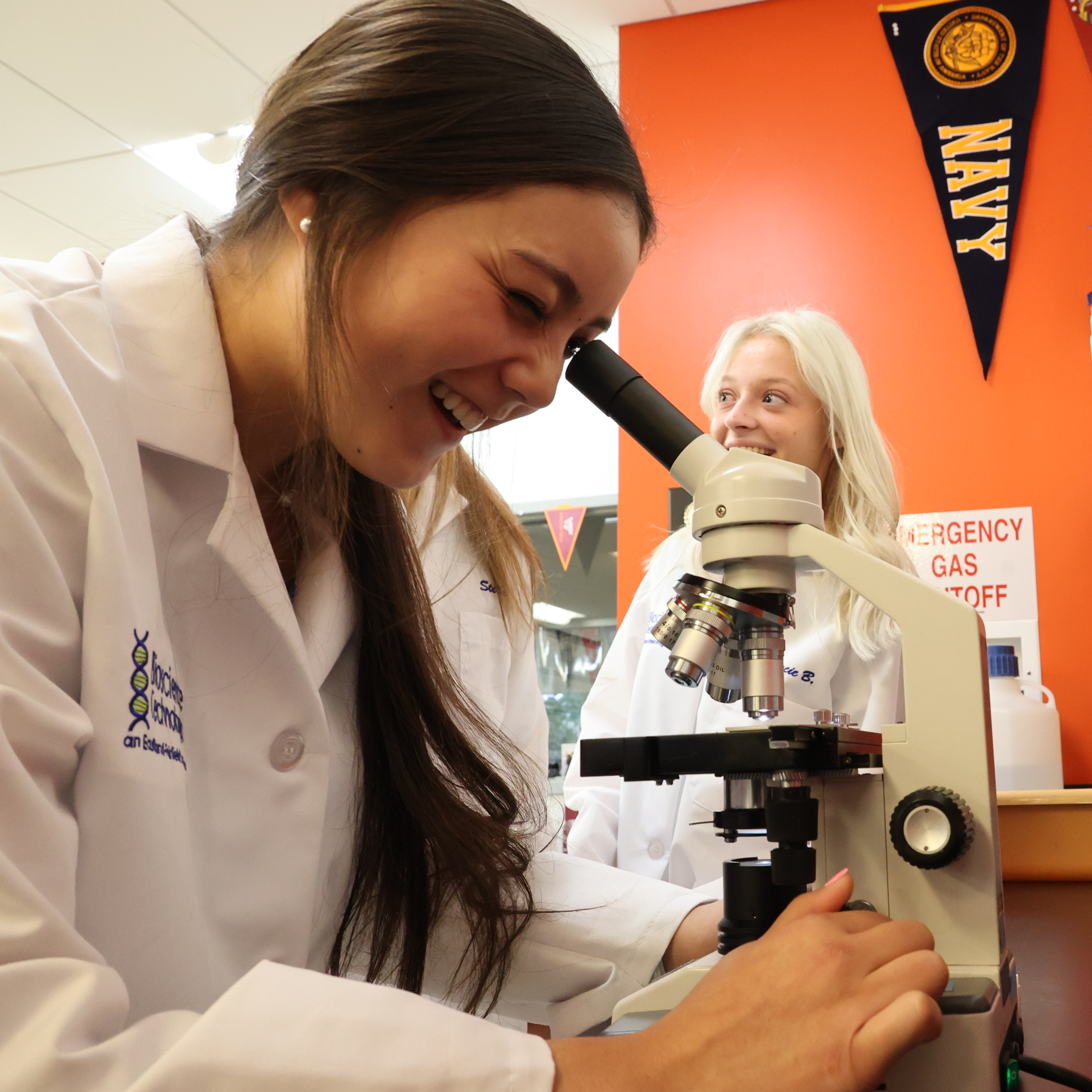 White, female student is smiling while looking through the lens of a tabletop microscope.