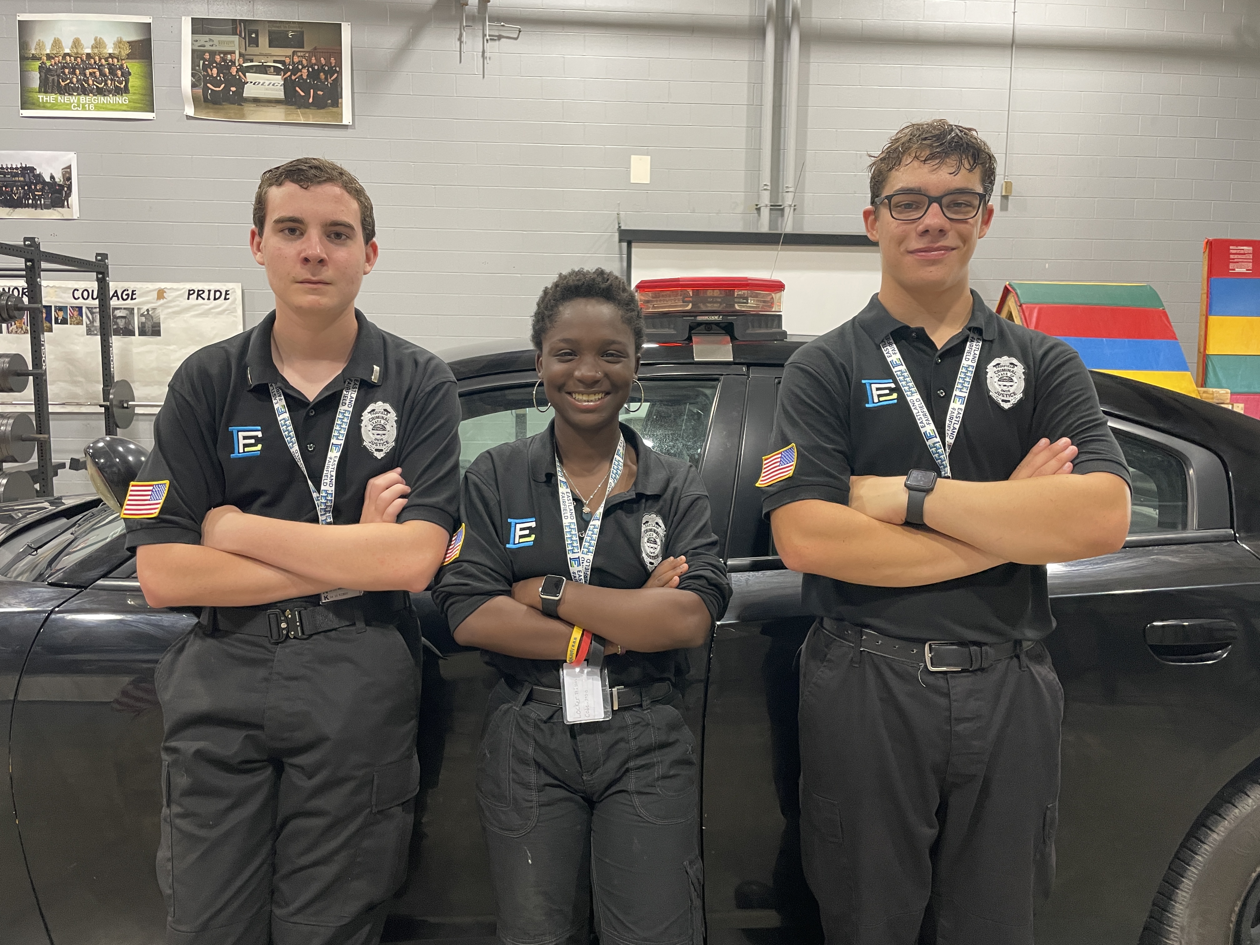 Two White male students flank a Black, female student. All have their arms crossed in front of them while they lean on a black police cruiser.