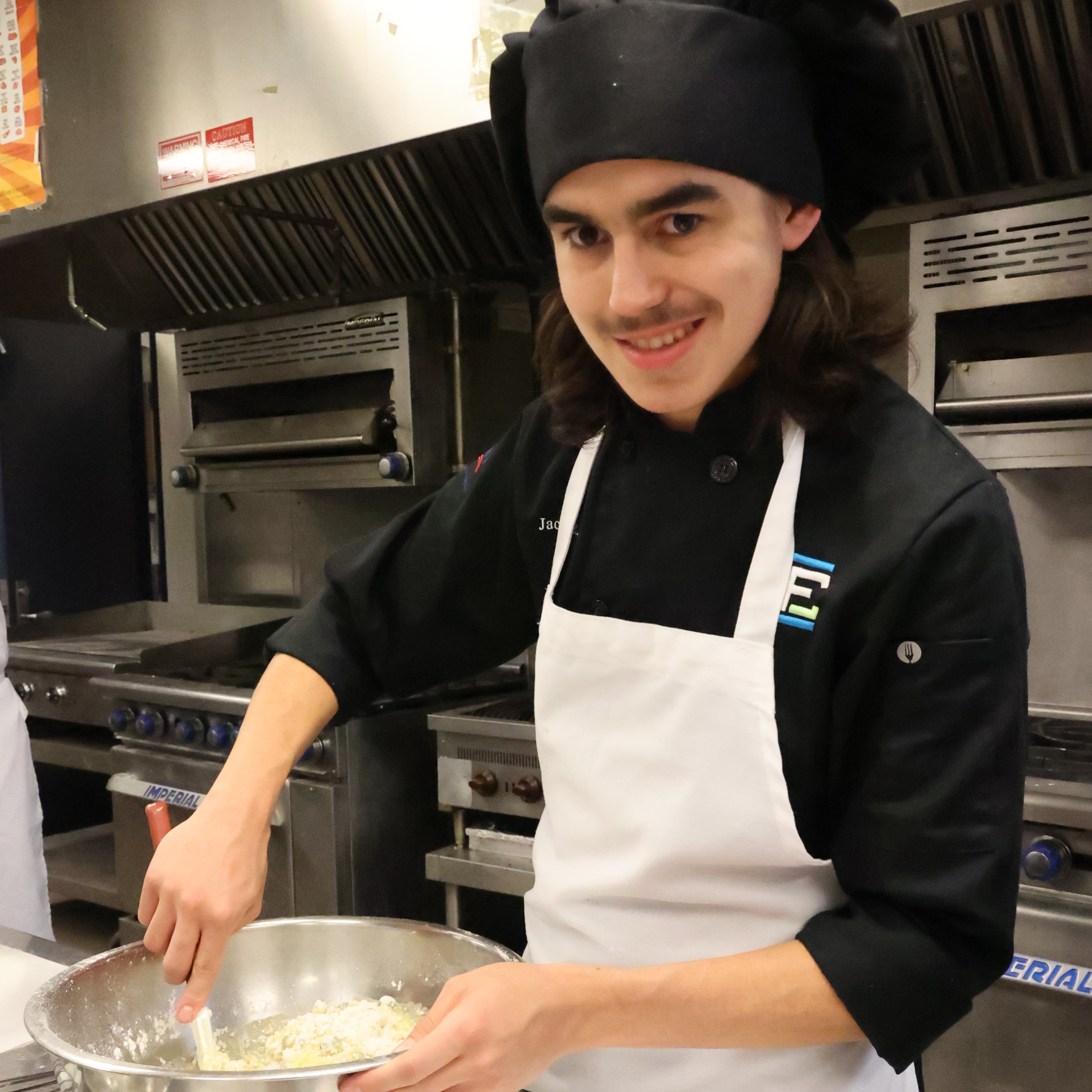 A male student with darker complexion wears his chef hat and apron while mixing ingredients in a mixing bowl.