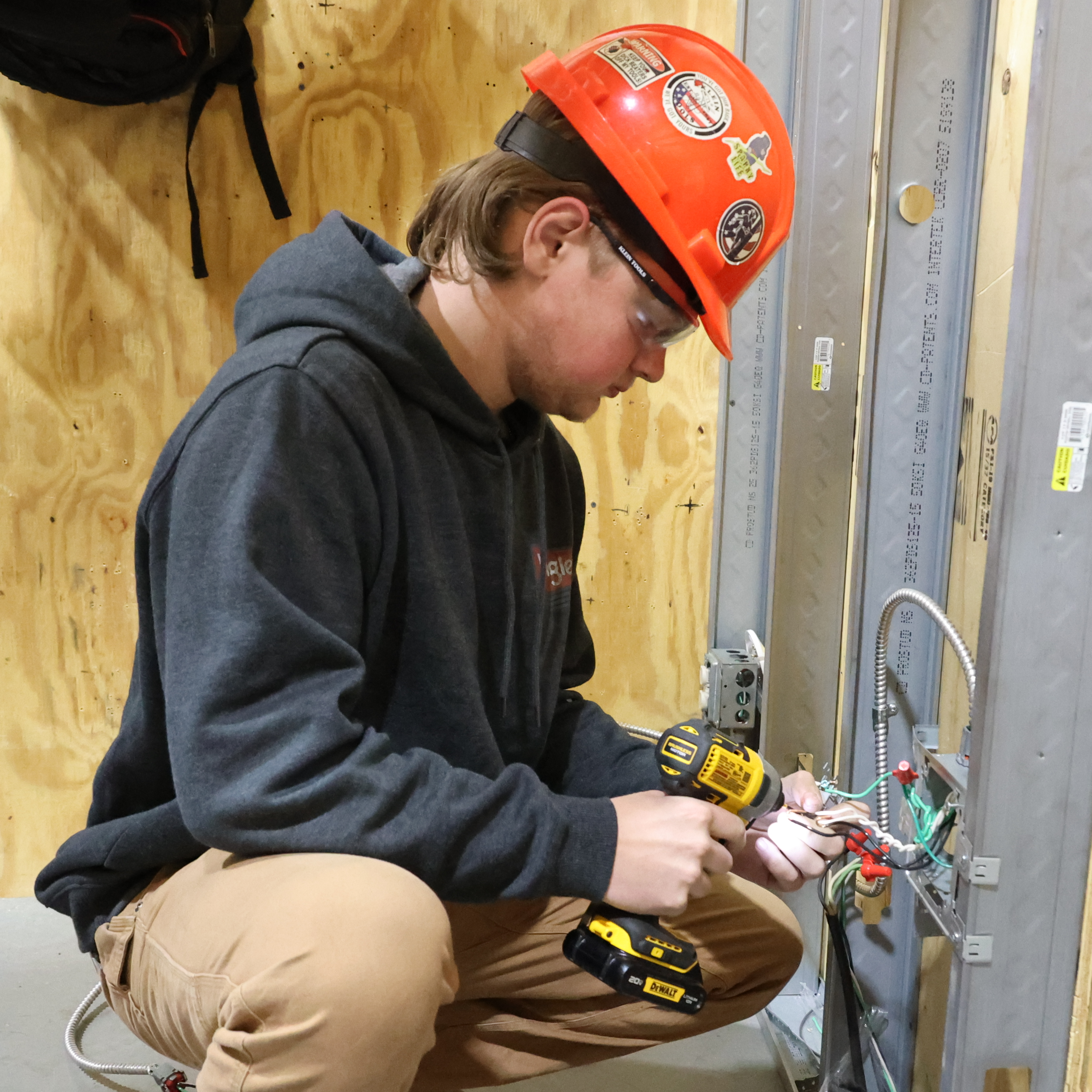 A White, male student wearing an orange hard hat holds a hand drill up to some loose cords on his wall work station.