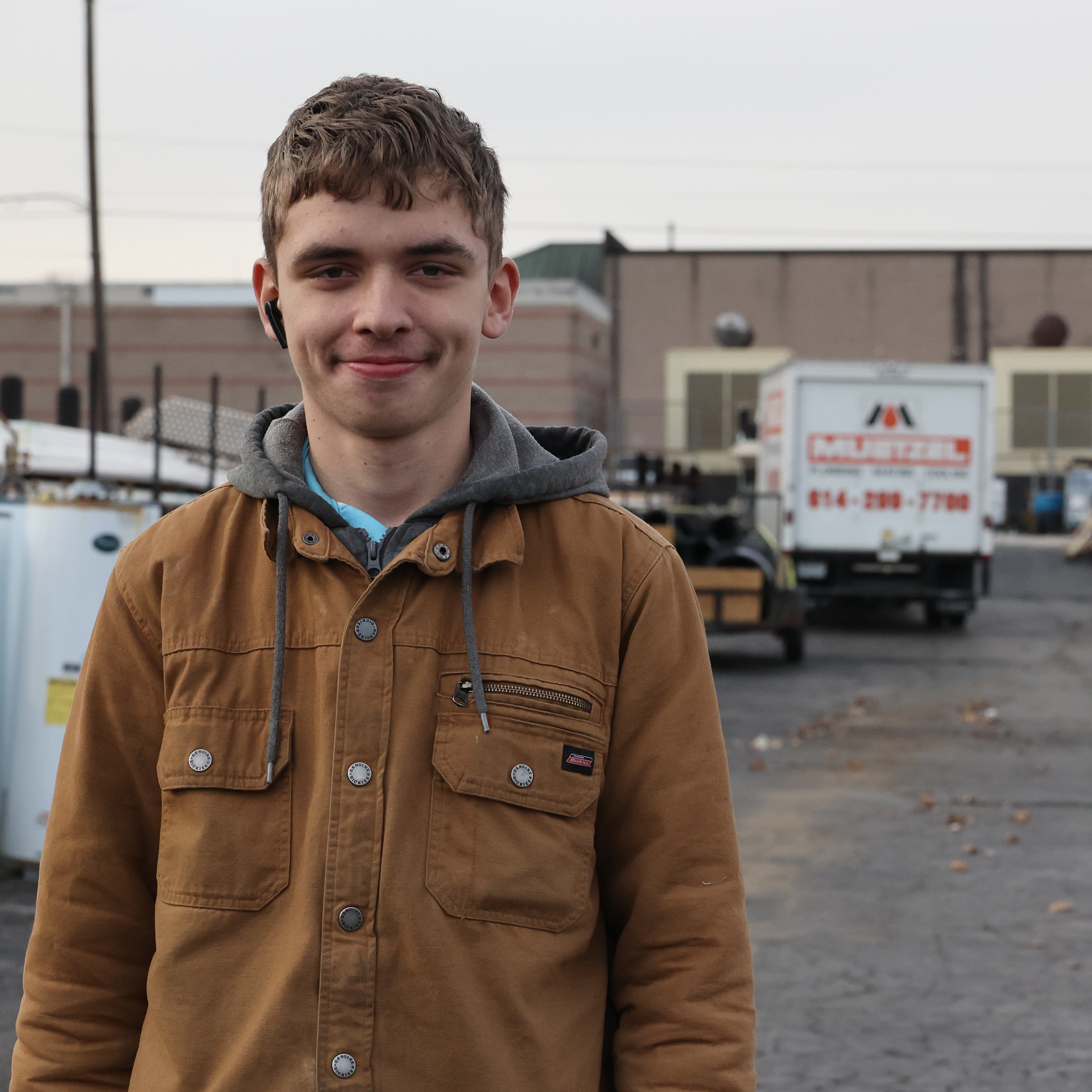 A White, male student stands in the parking lot of a warehouse wearing a brown winter jacket with cargo trucks parked behind him.