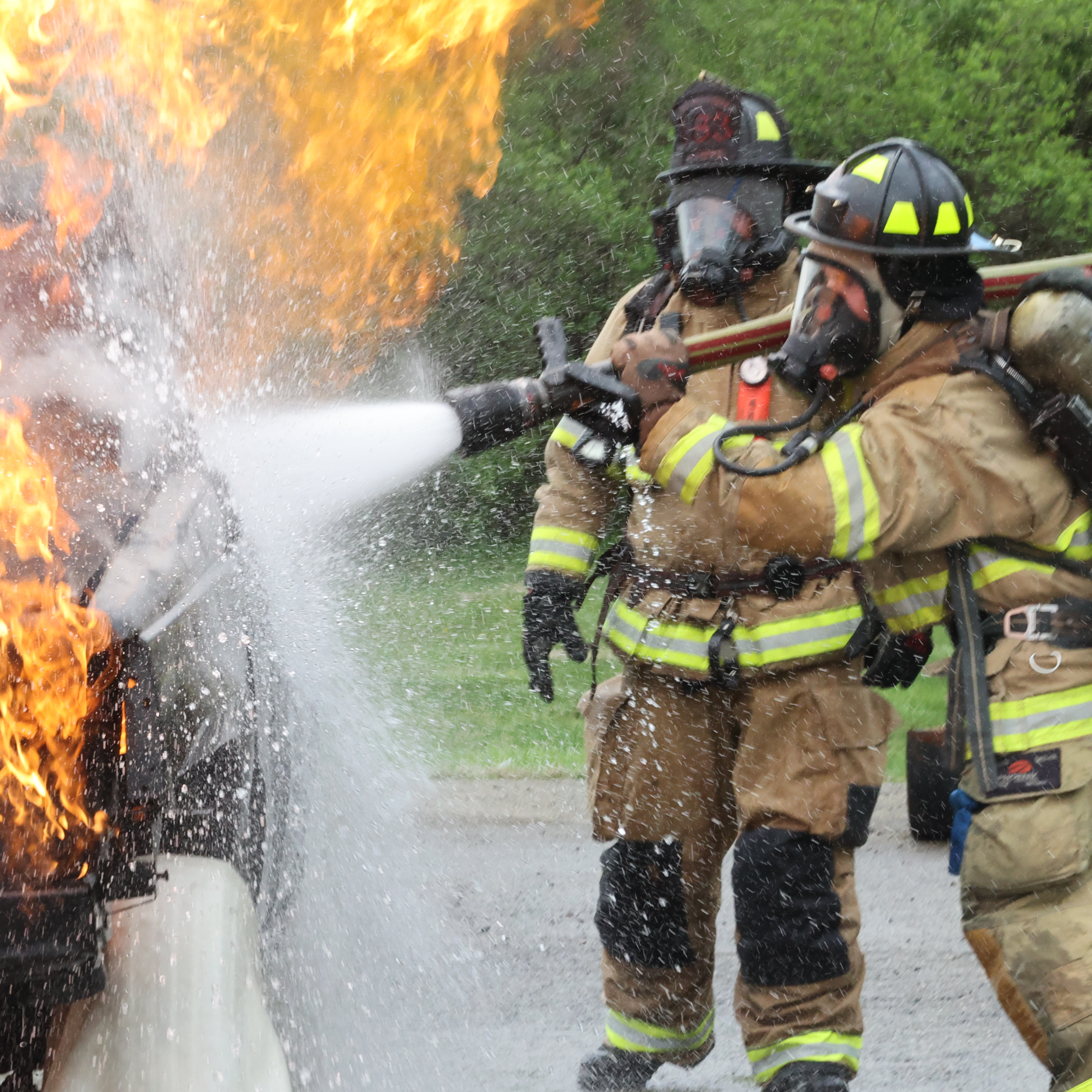 Firefighting students are holding a hose and spraying water on a car fire.