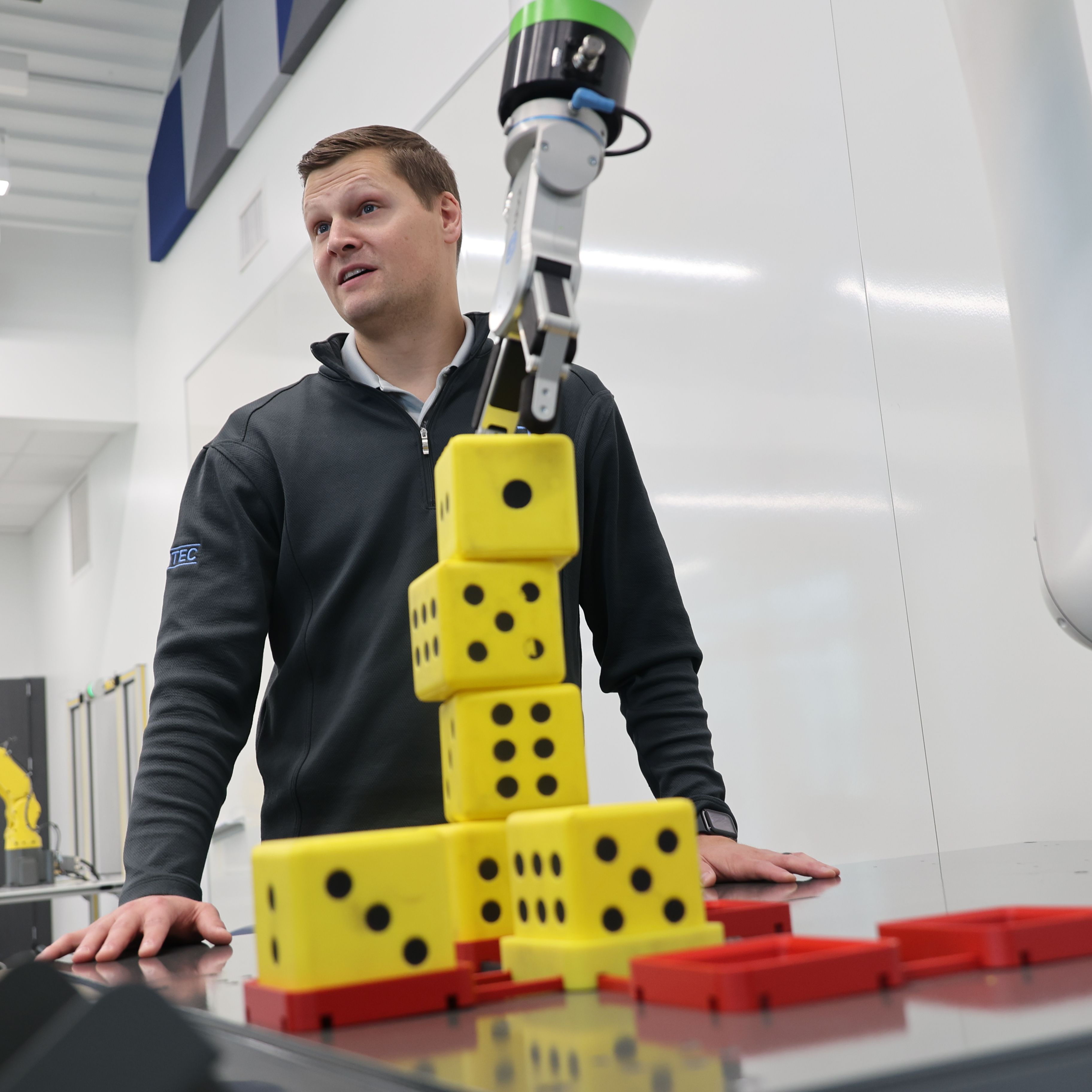 A White male models how a robotic arm can pick up a stack of yellow, foam dice