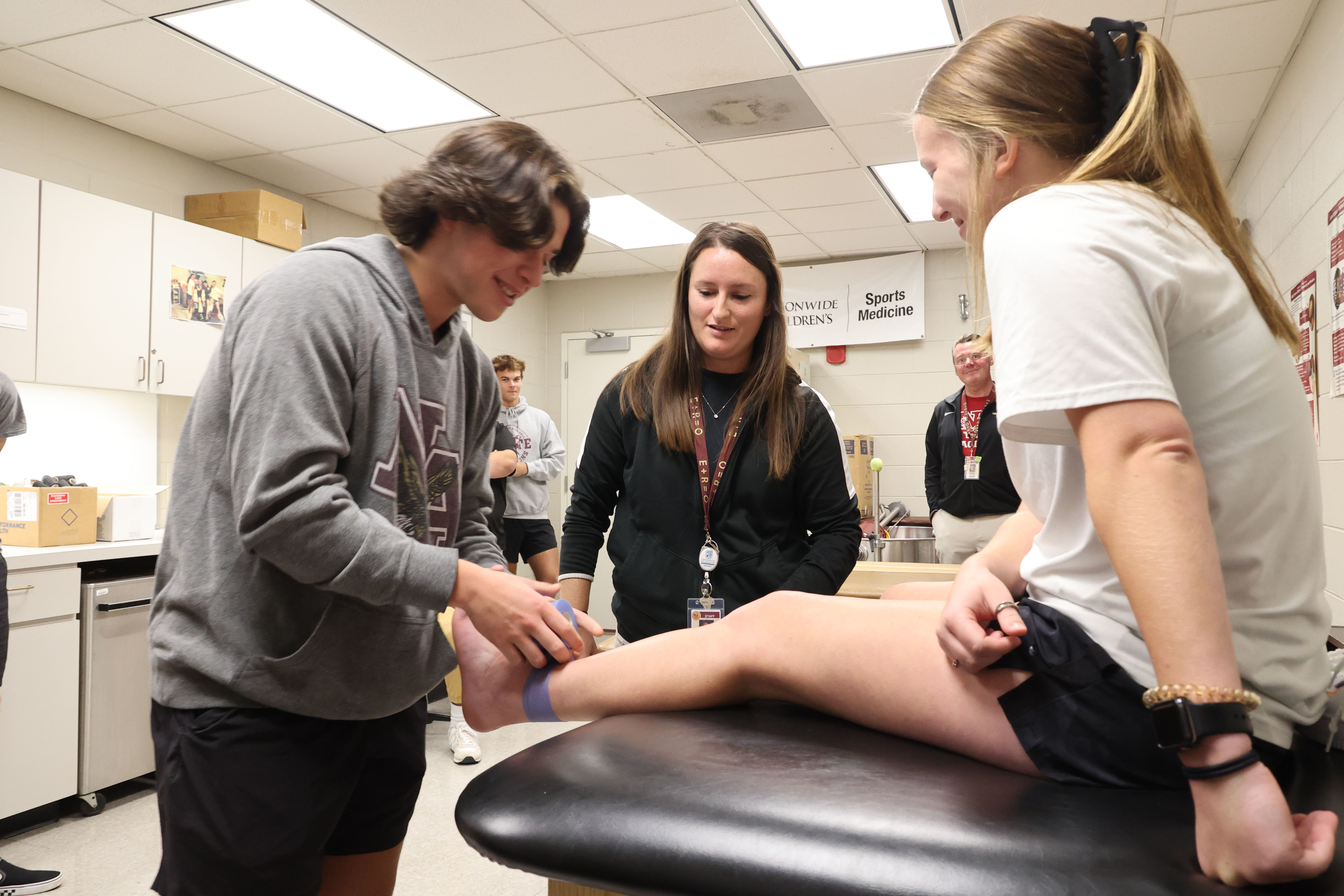 A White male student begins applying medical wrap to the ankle of a White, female student under the supervision on their instructor.